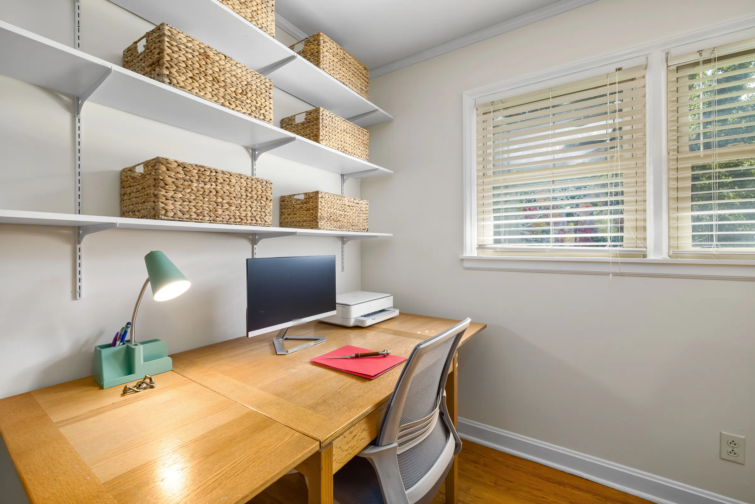 A small bright room set up as an office, with a wood desk, a monitor and a green lamp under white wall shelves holding woven baskets