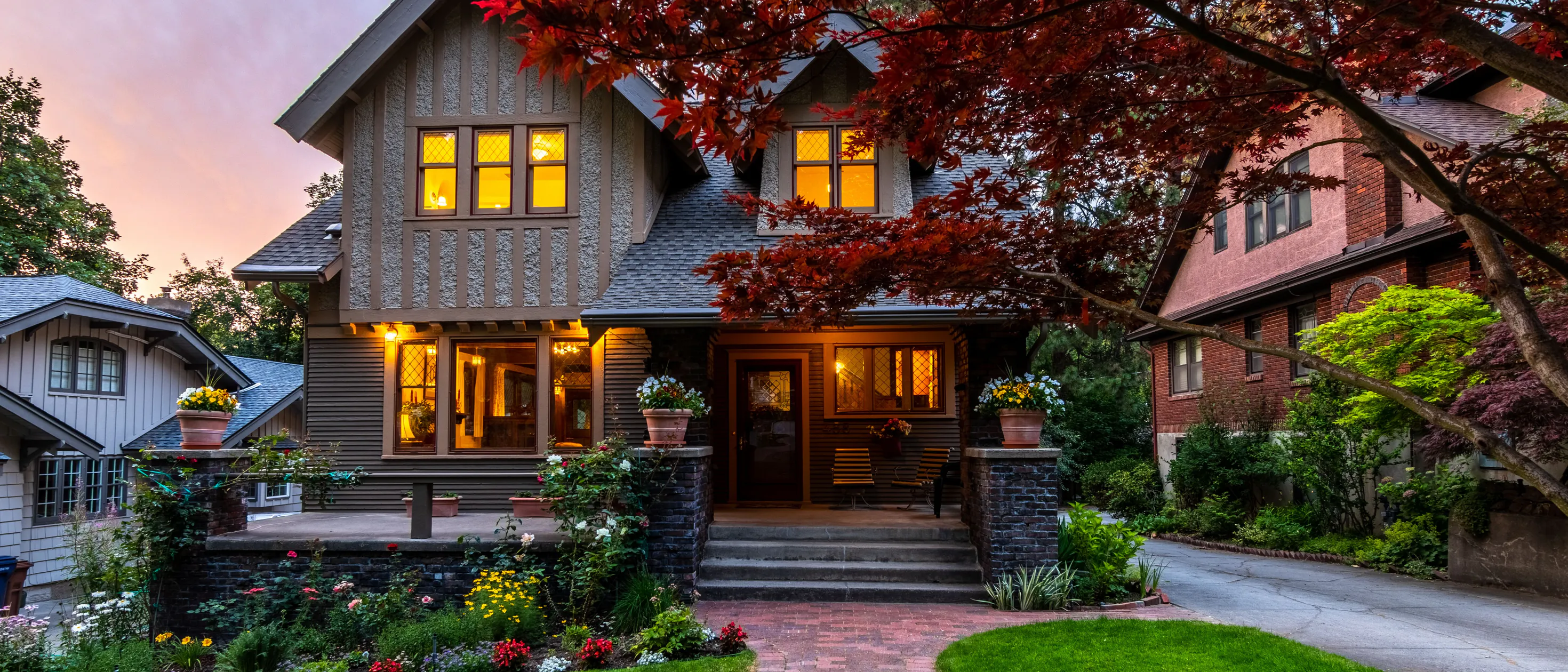 A Craftsman with lit windows at dusk behind a brick path, a lawn and a red Japanese maple
