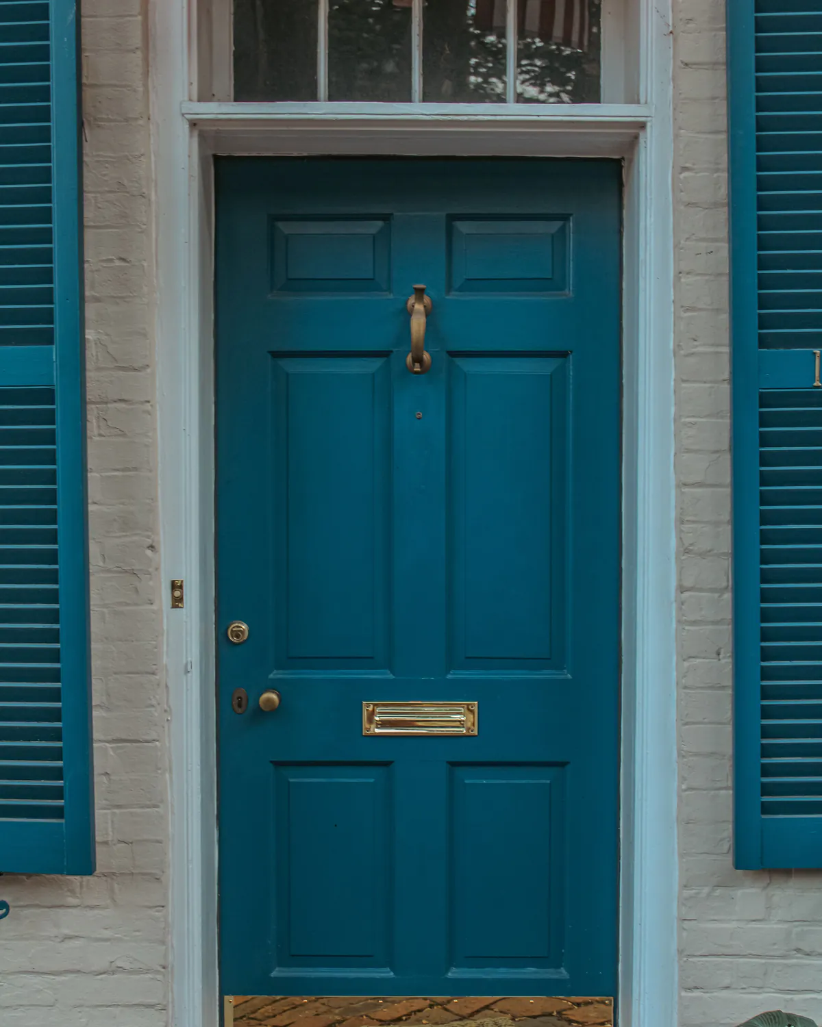 A six-panel front door in a deep teal gloss with a brass knocker and letter plate, between teal louvred shutters