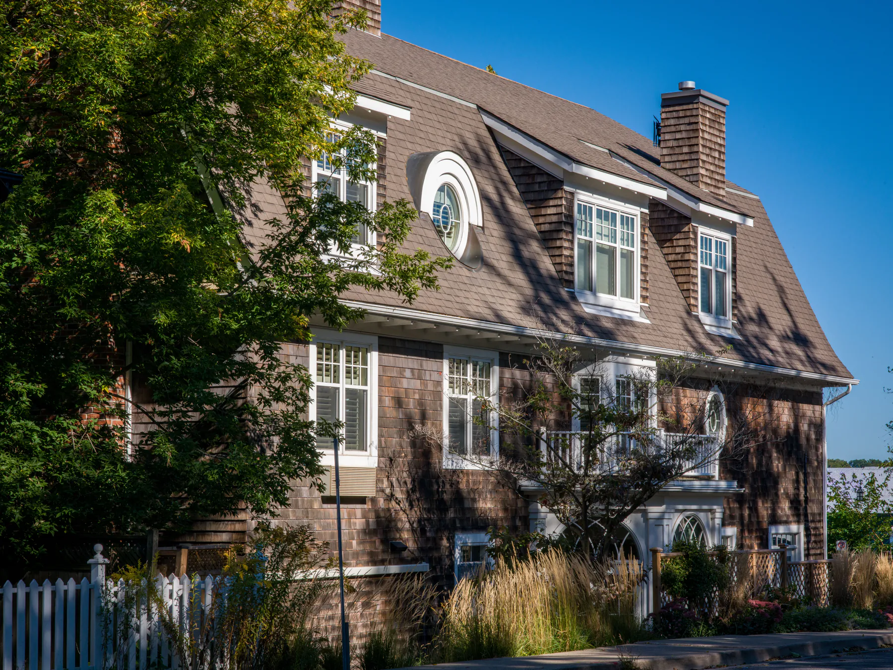 A brown-shingled house with white-trimmed windows, a round attic window and a picket fence under a blue sky