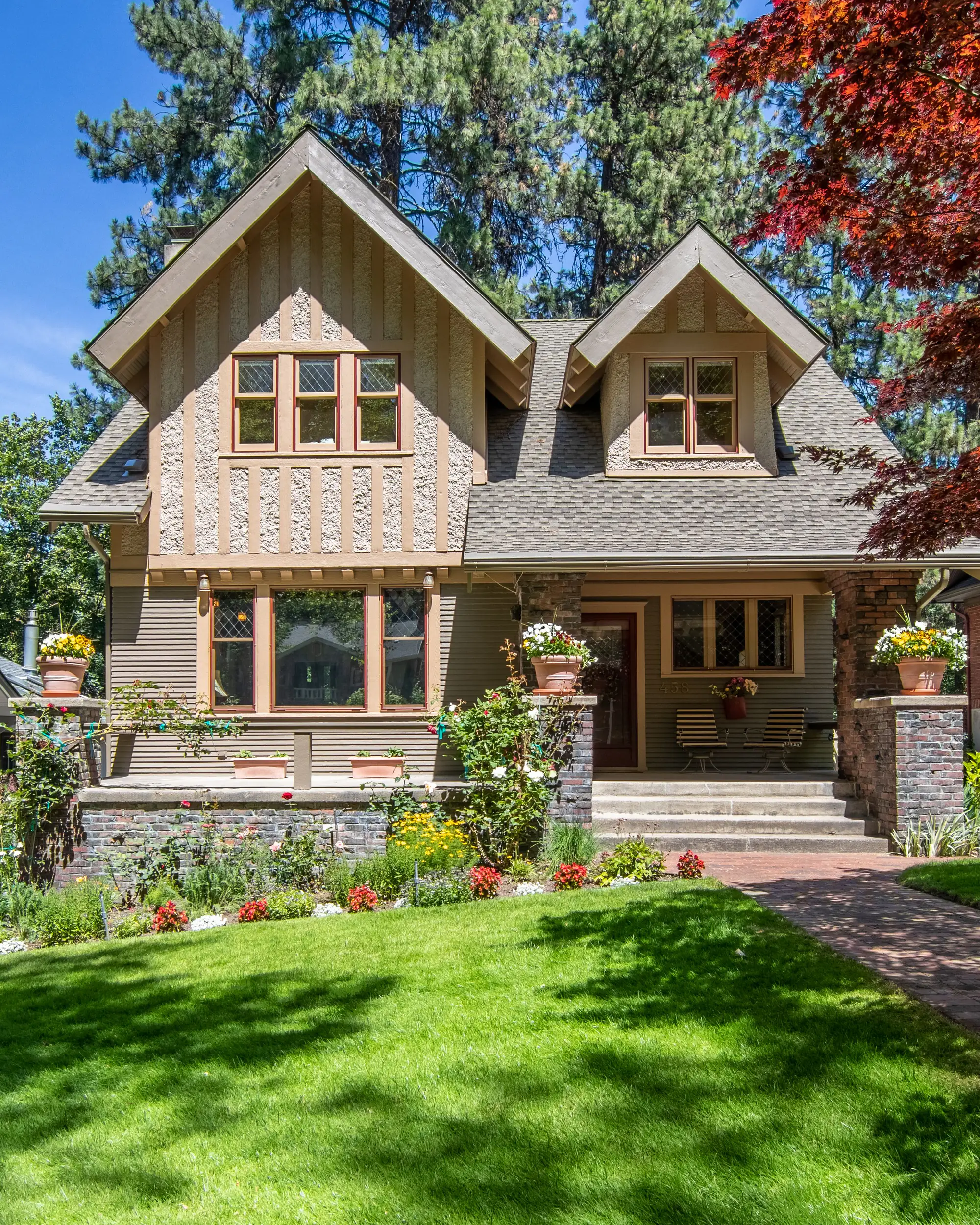 A two-storey Craftsman with tan shingle siding, brown trim and twin front gables above a sunlit lawn