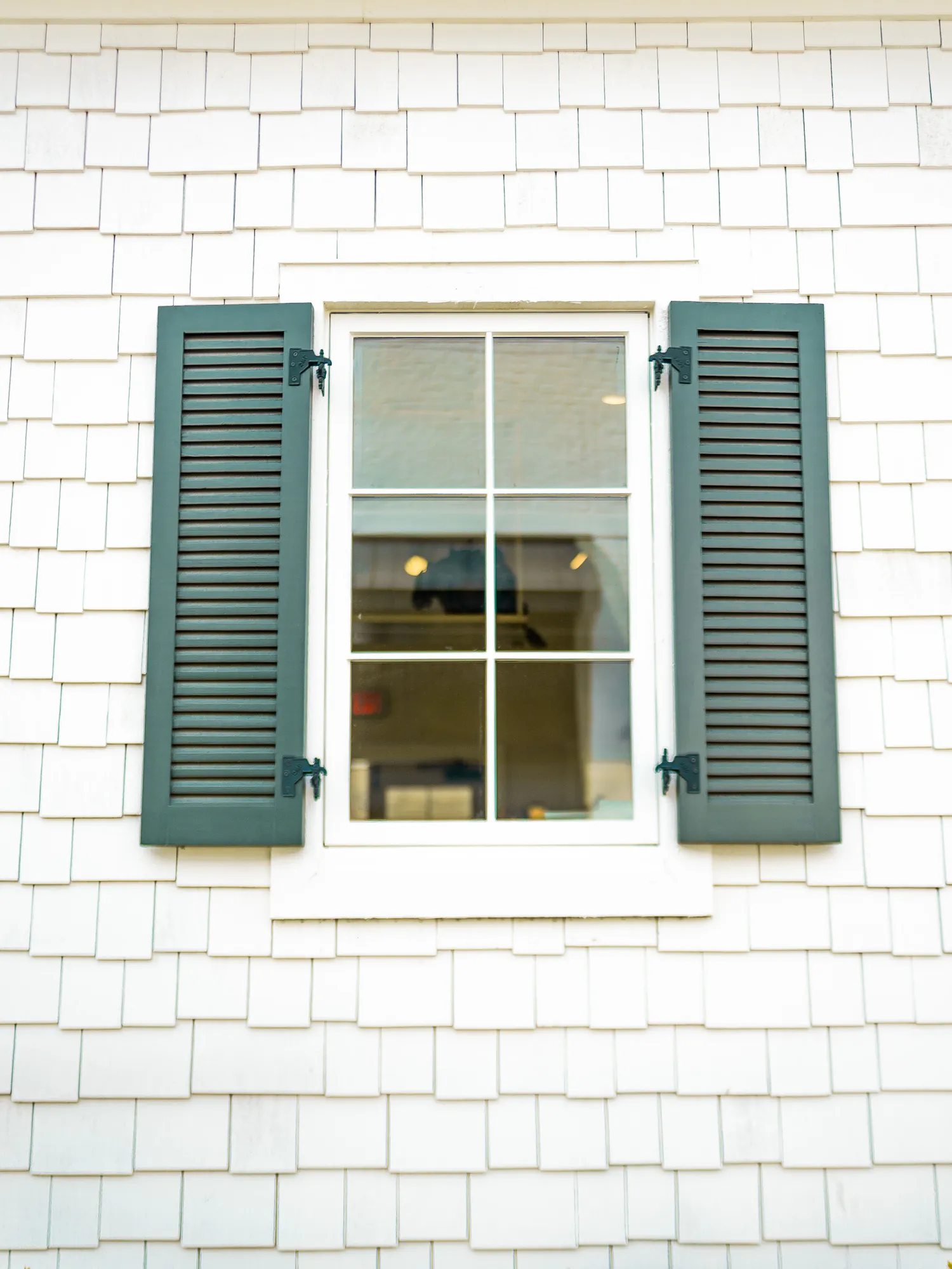 A white shingle wall with a six-light window between dark green louvred shutters, under a painted eave