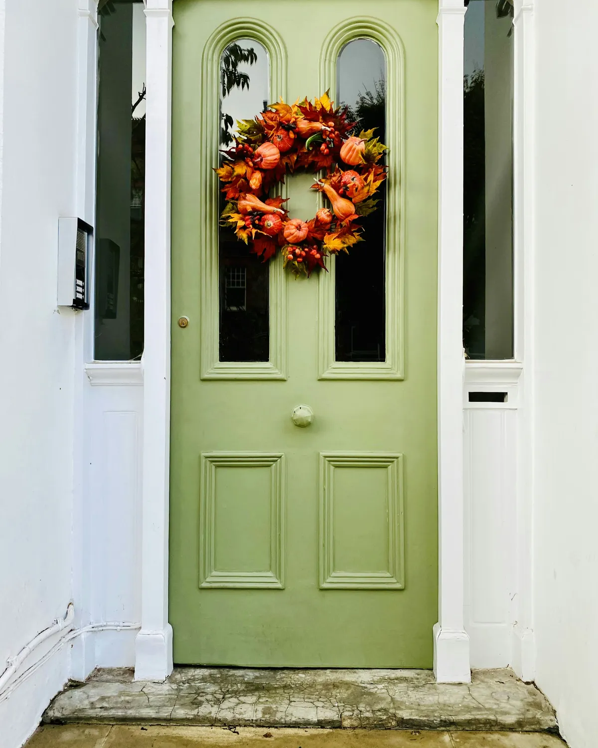 A sage green front door with arched glass panes and an autumn wreath, between white sidelights