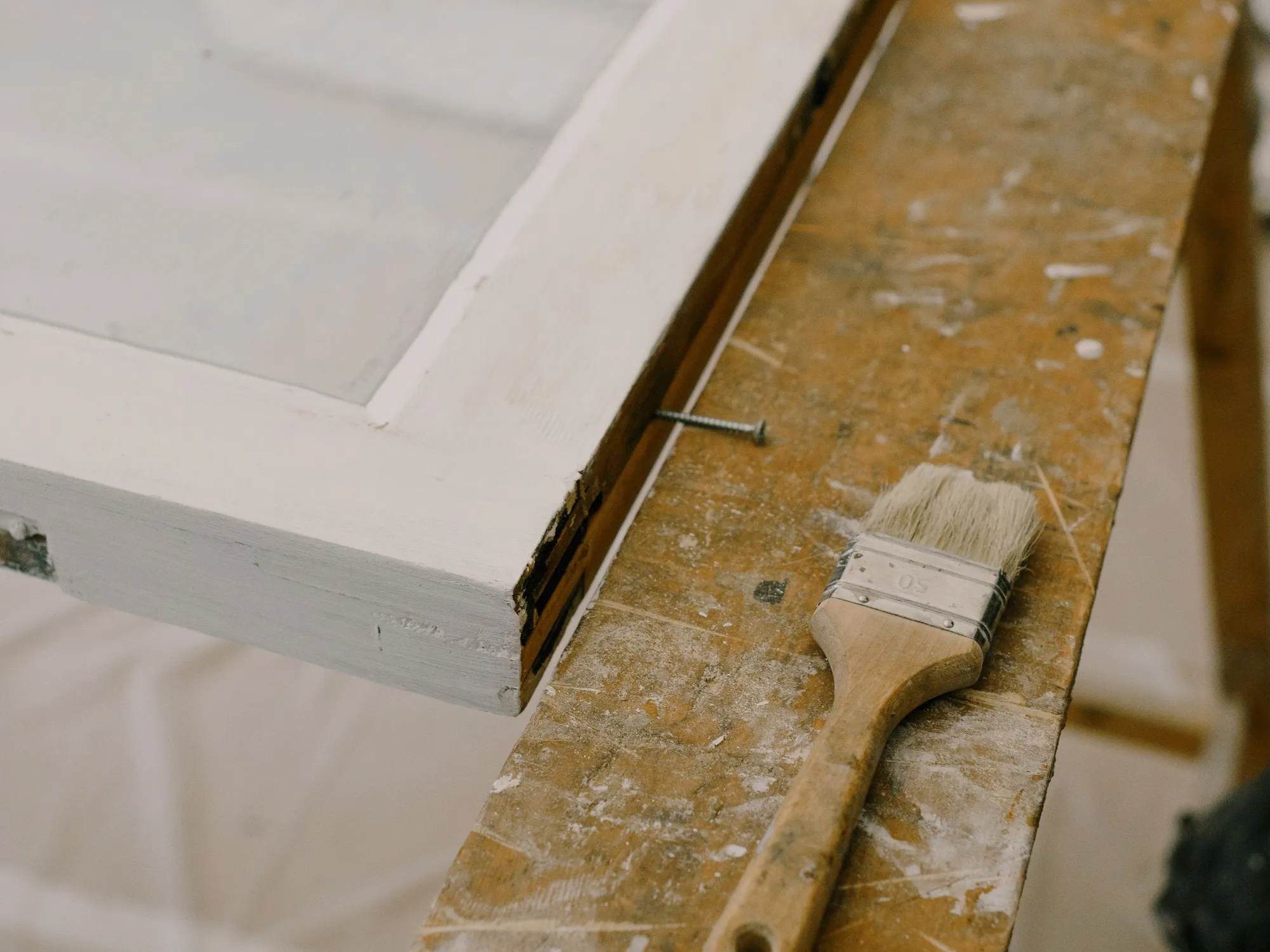 A freshly painted white window sash on a paint-spattered workbench beside a brush