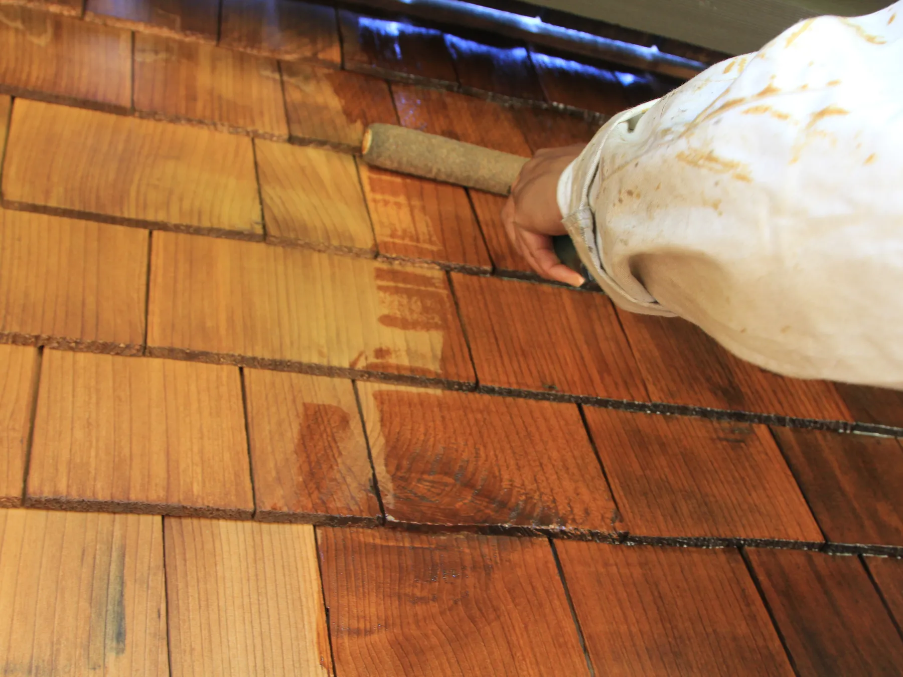 A sleeved arm rolling stain onto cedar shingles under an eave