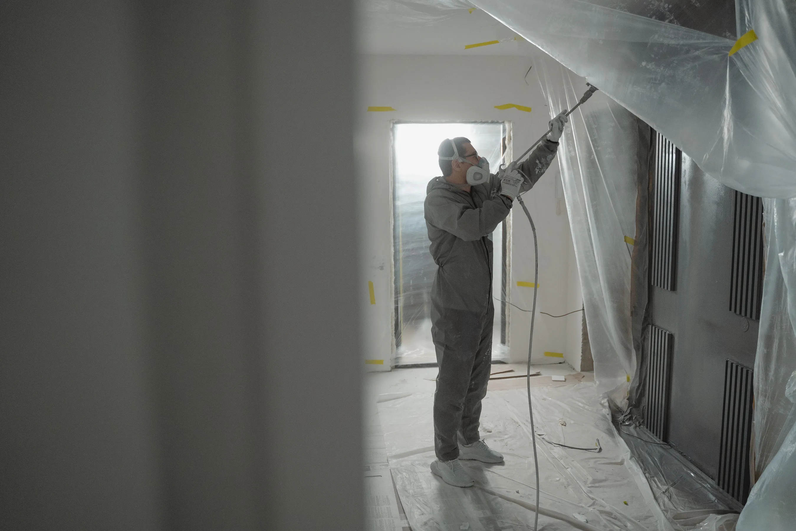 A painter in a hooded suit and respirator spraying a wall in a room masked off with plastic sheeting