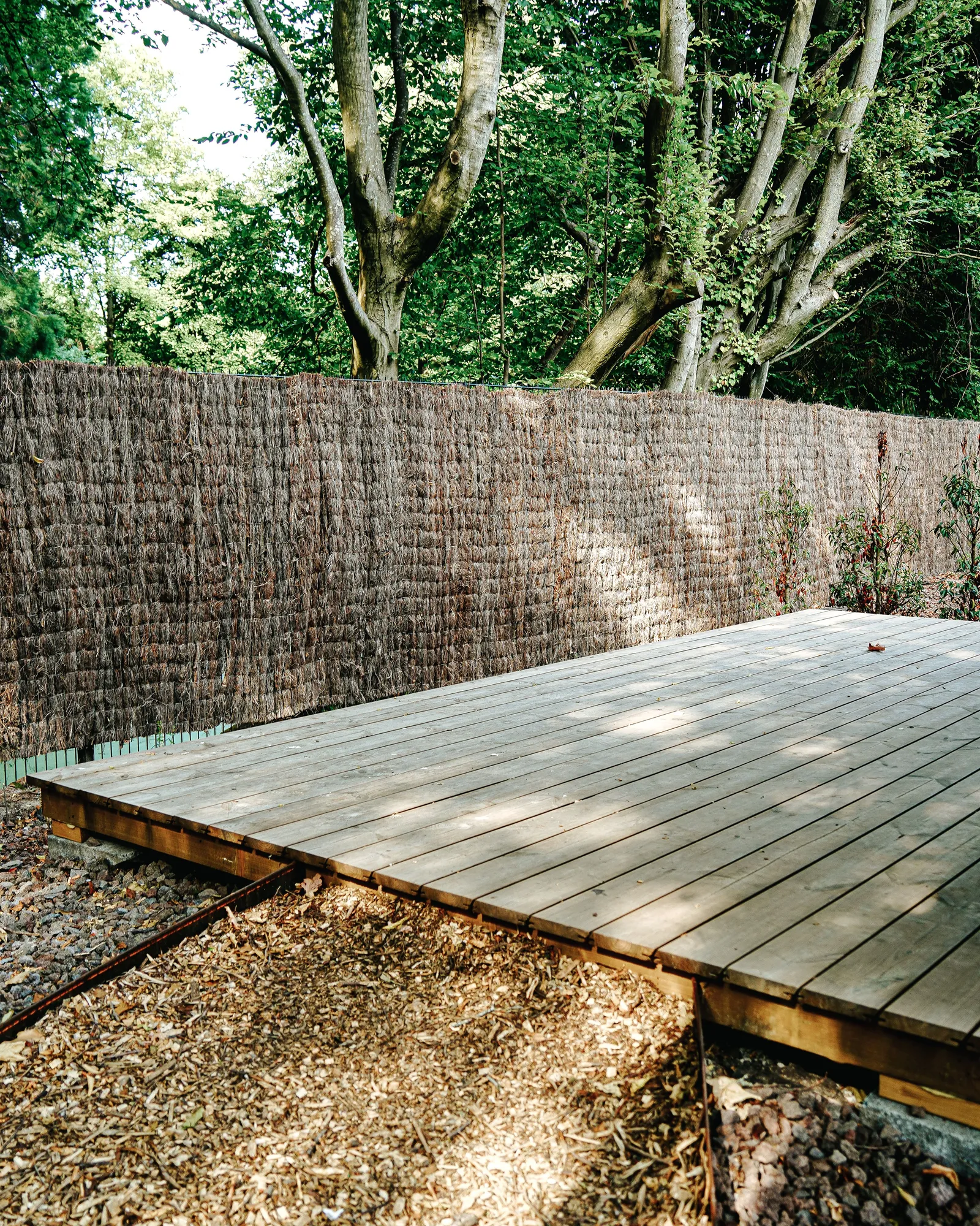 A low wood deck raised over bare gravel and dry ground in front of a reed fence and trees