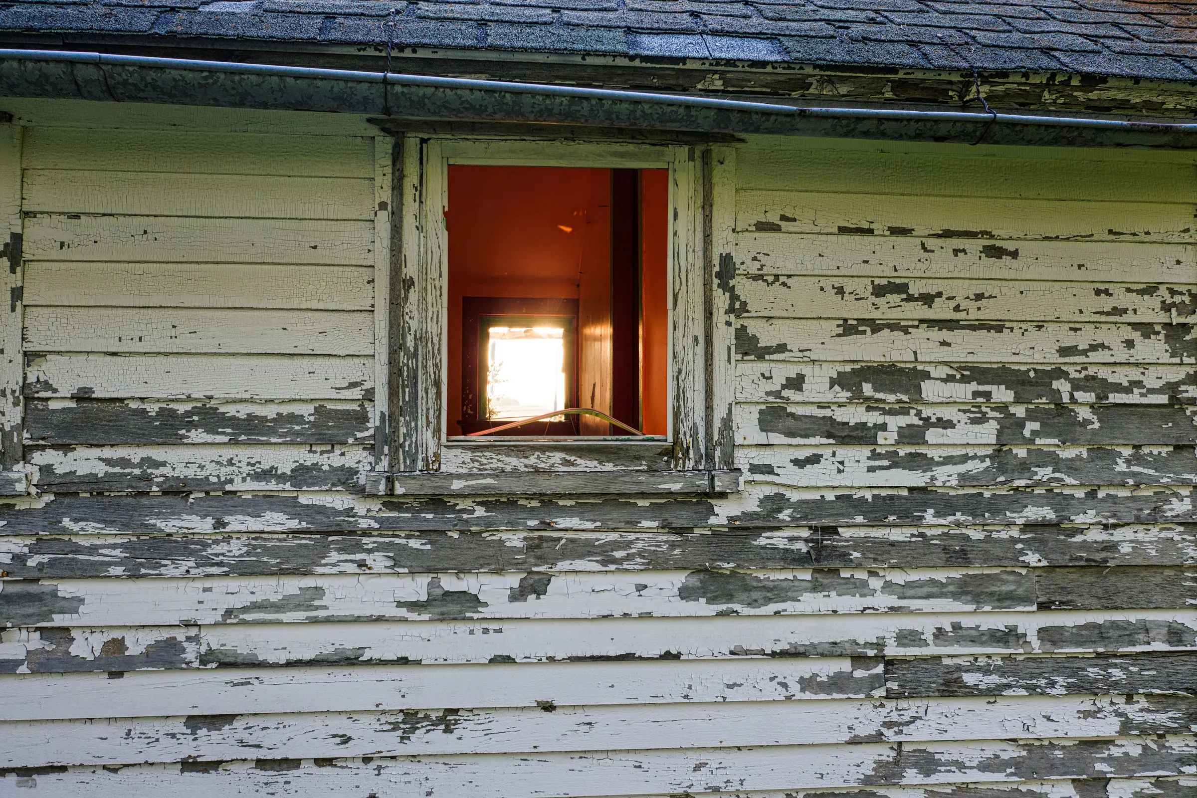 Old wood lap siding with its white paint flaking away around a small window under the eave