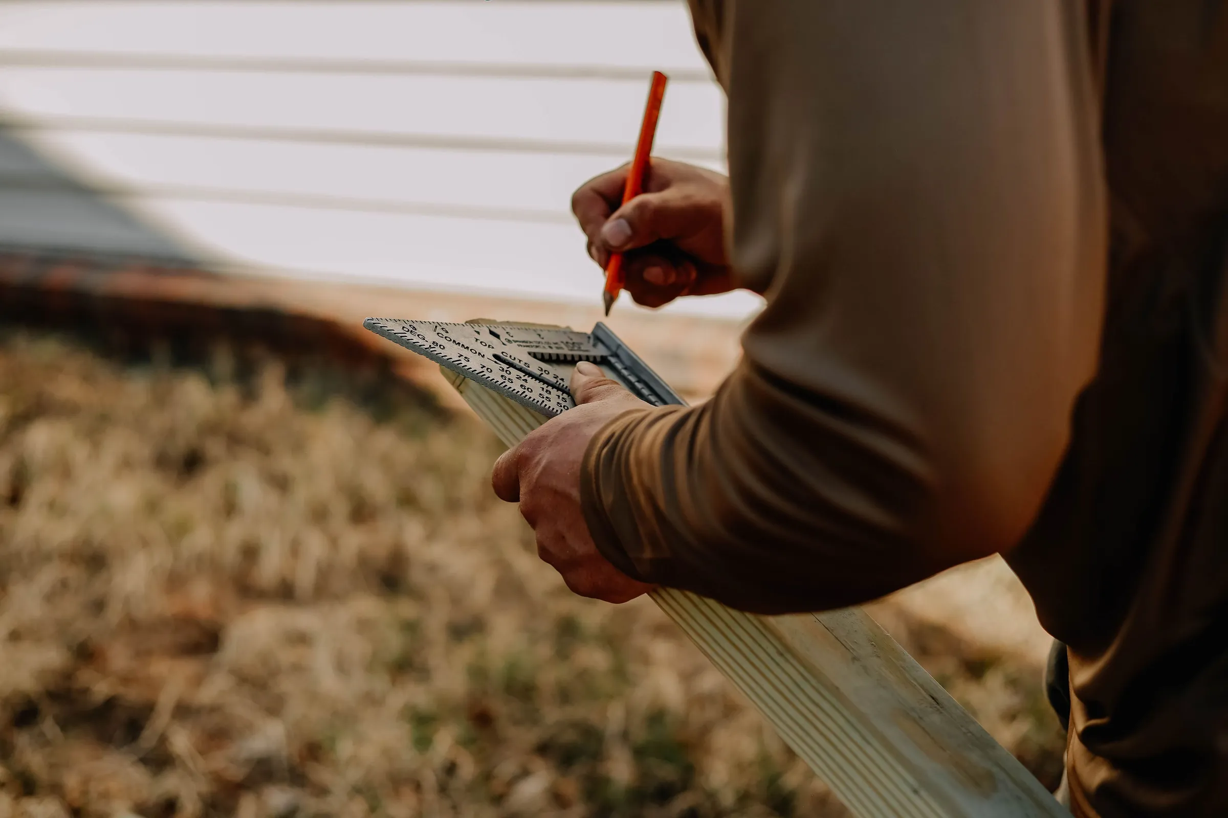 A hand marking a board with a pencil and a speed square, white lap siding behind