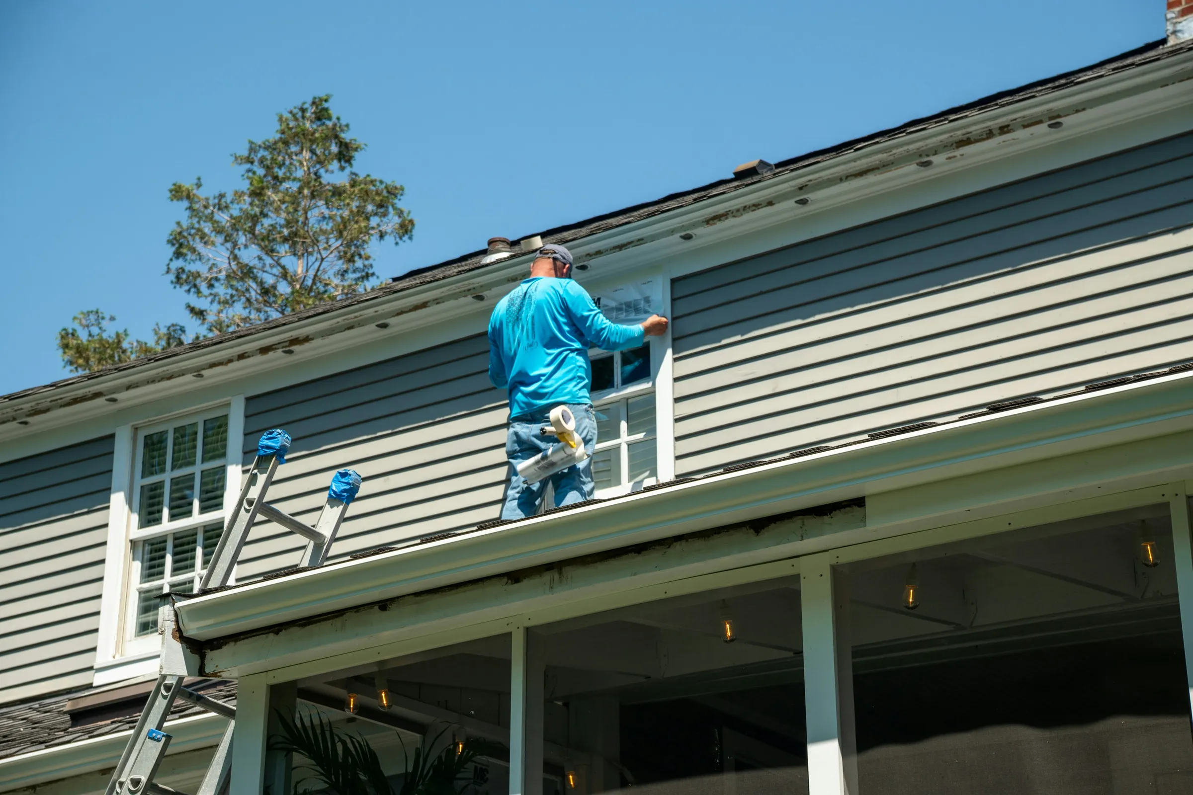 A worker seen from behind on a porch roof, taping around an upstairs window in grey lap siding