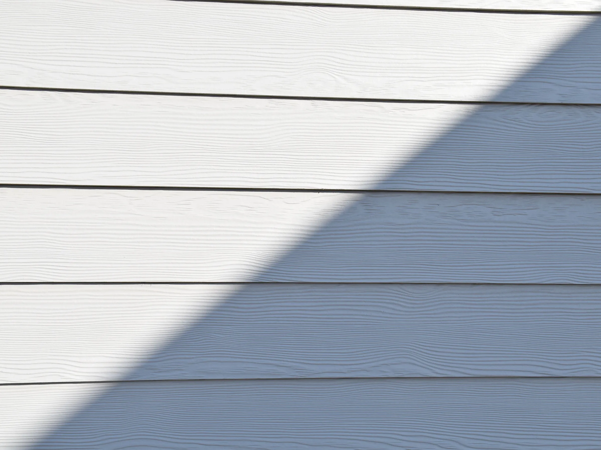 A run of white wood-grain lap siding, half in sunlight and half under a sharp diagonal shadow