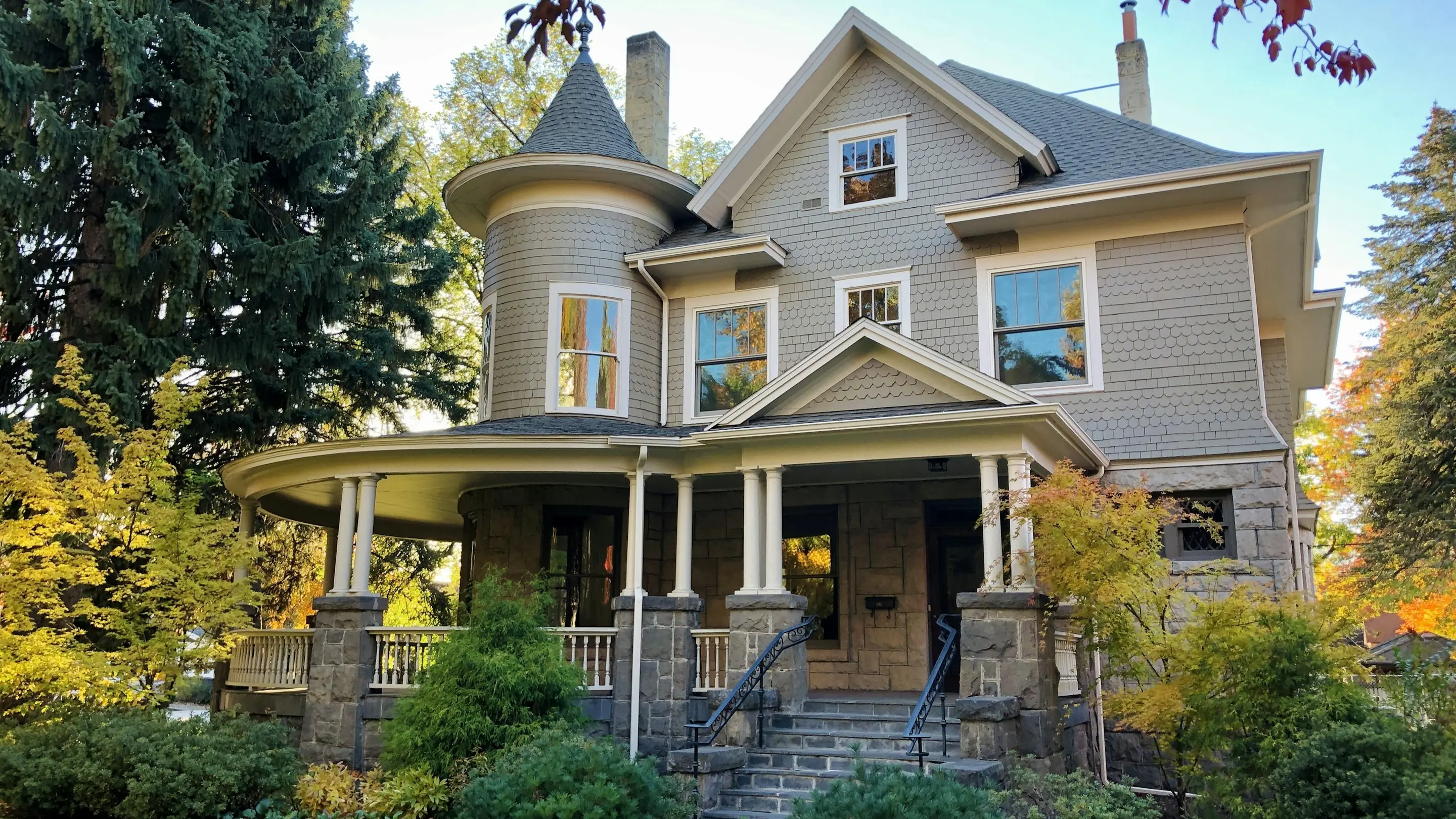 A grey shingled Queen Anne house with a round corner turret and a wraparound porch under autumn trees