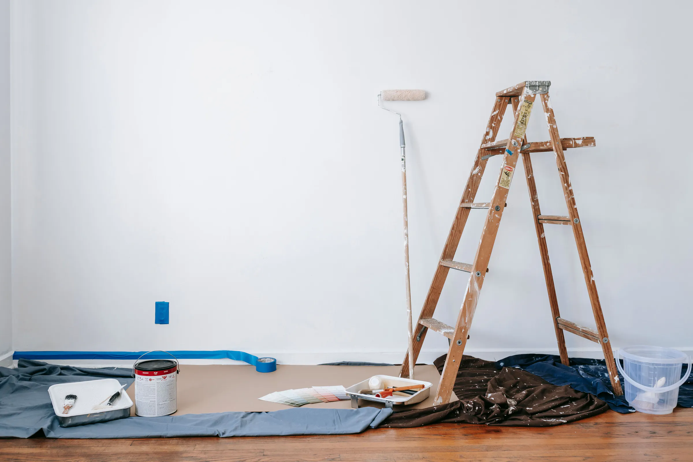 A white room prepared for painting: a wooden stepladder, a roller, paint trays and drop cloths taped along the baseboard