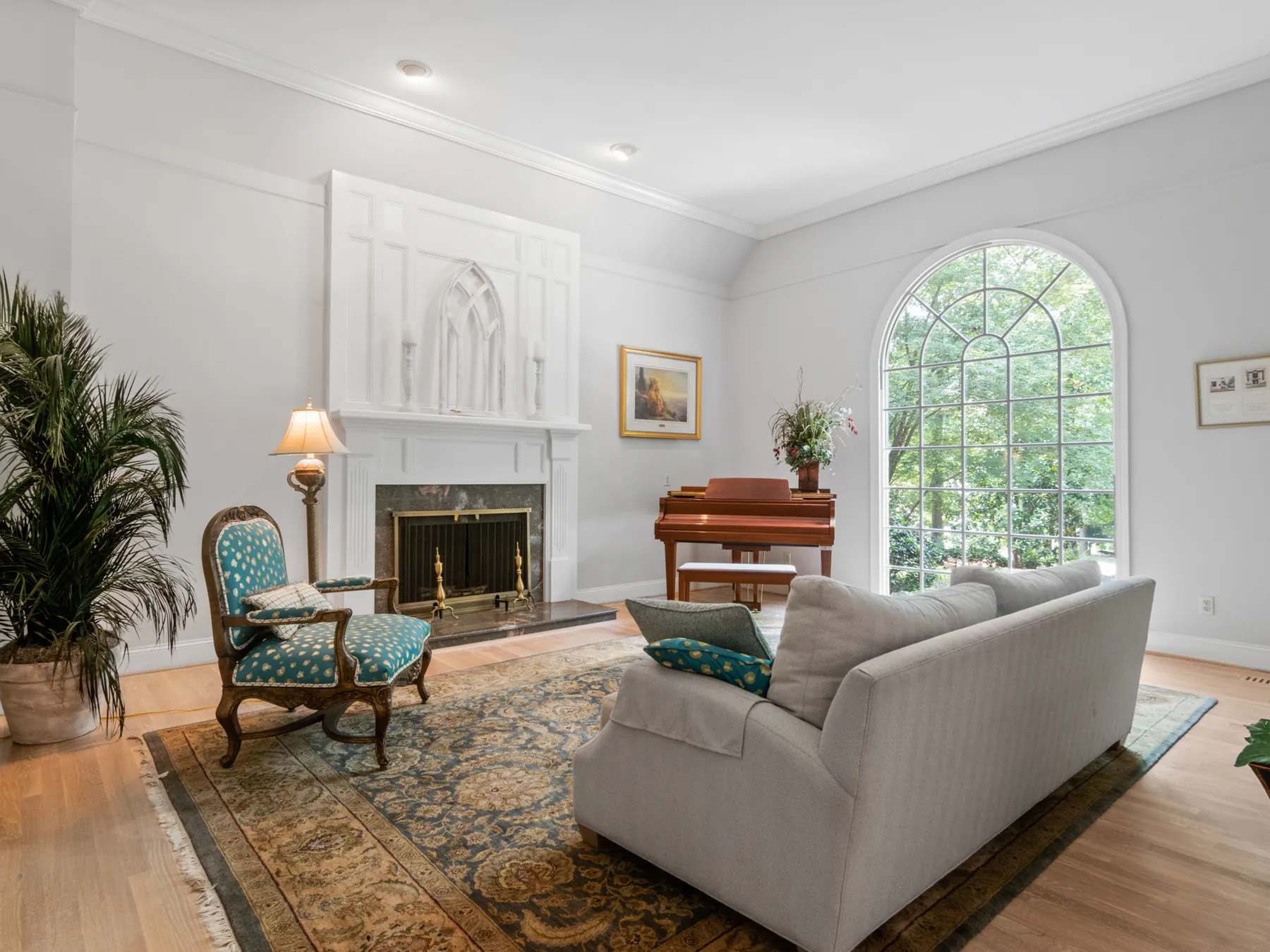 A bright living room with pale grey walls, a white fireplace surround, an arched window and a piano