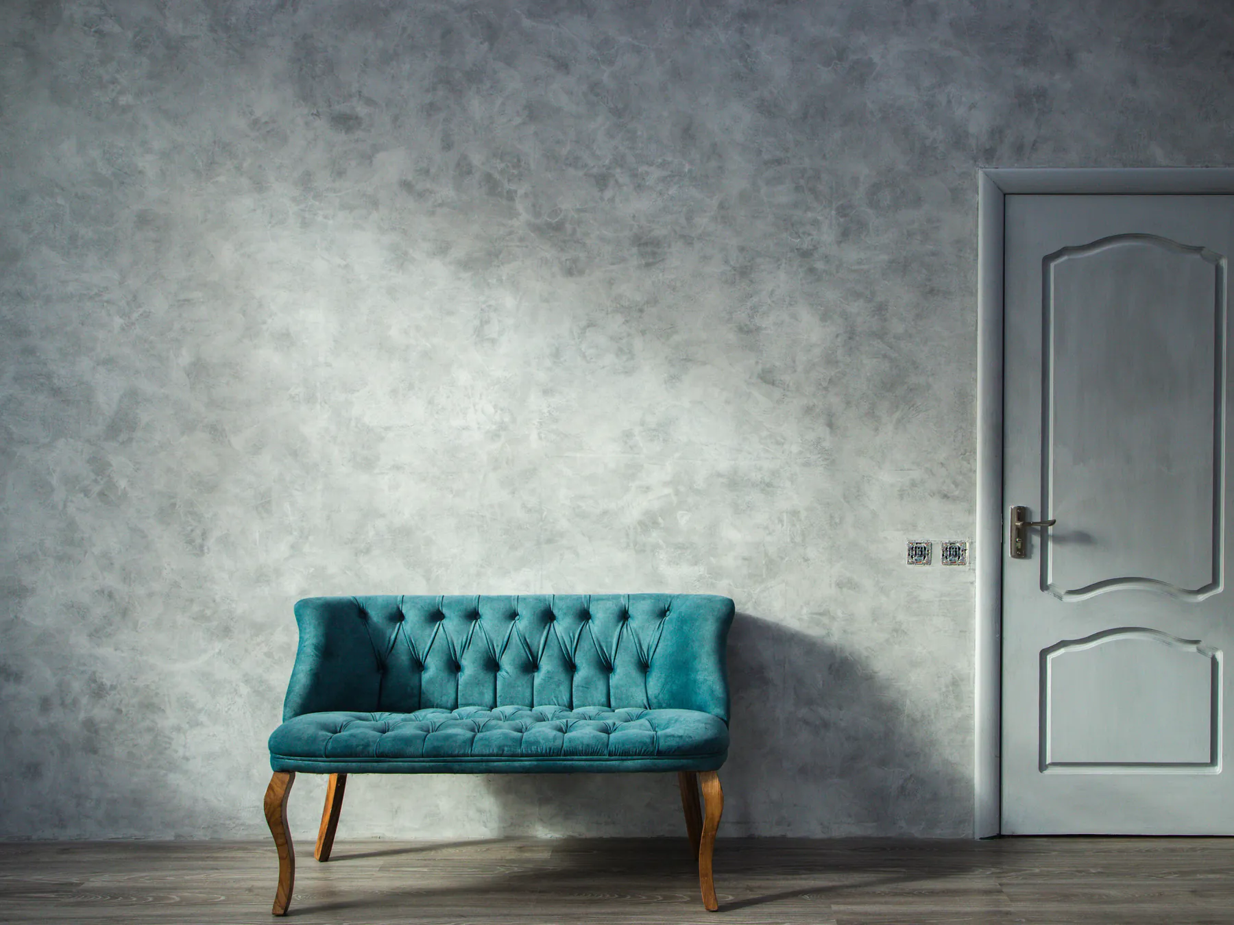A teal tufted settee against a mottled grey plaster wall beside a panelled door