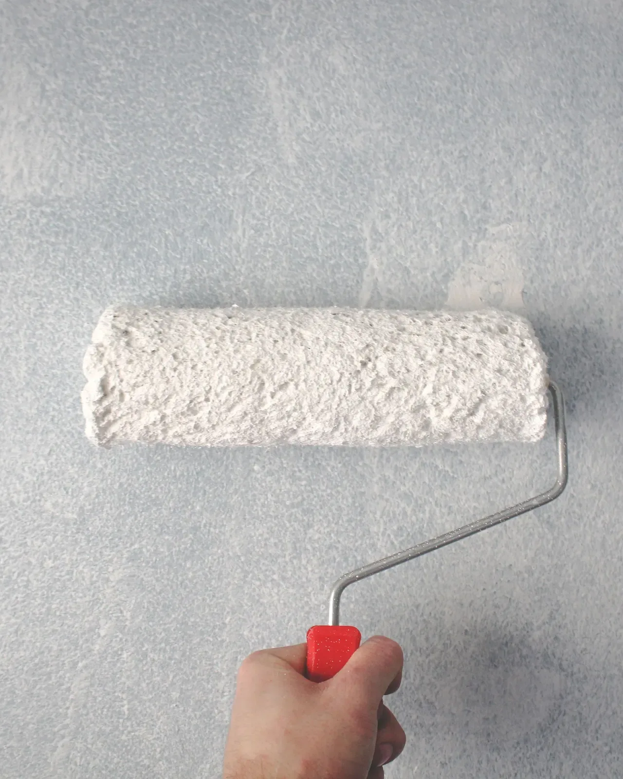 A hand rolling white paint over a grey wall with a red handled roller
