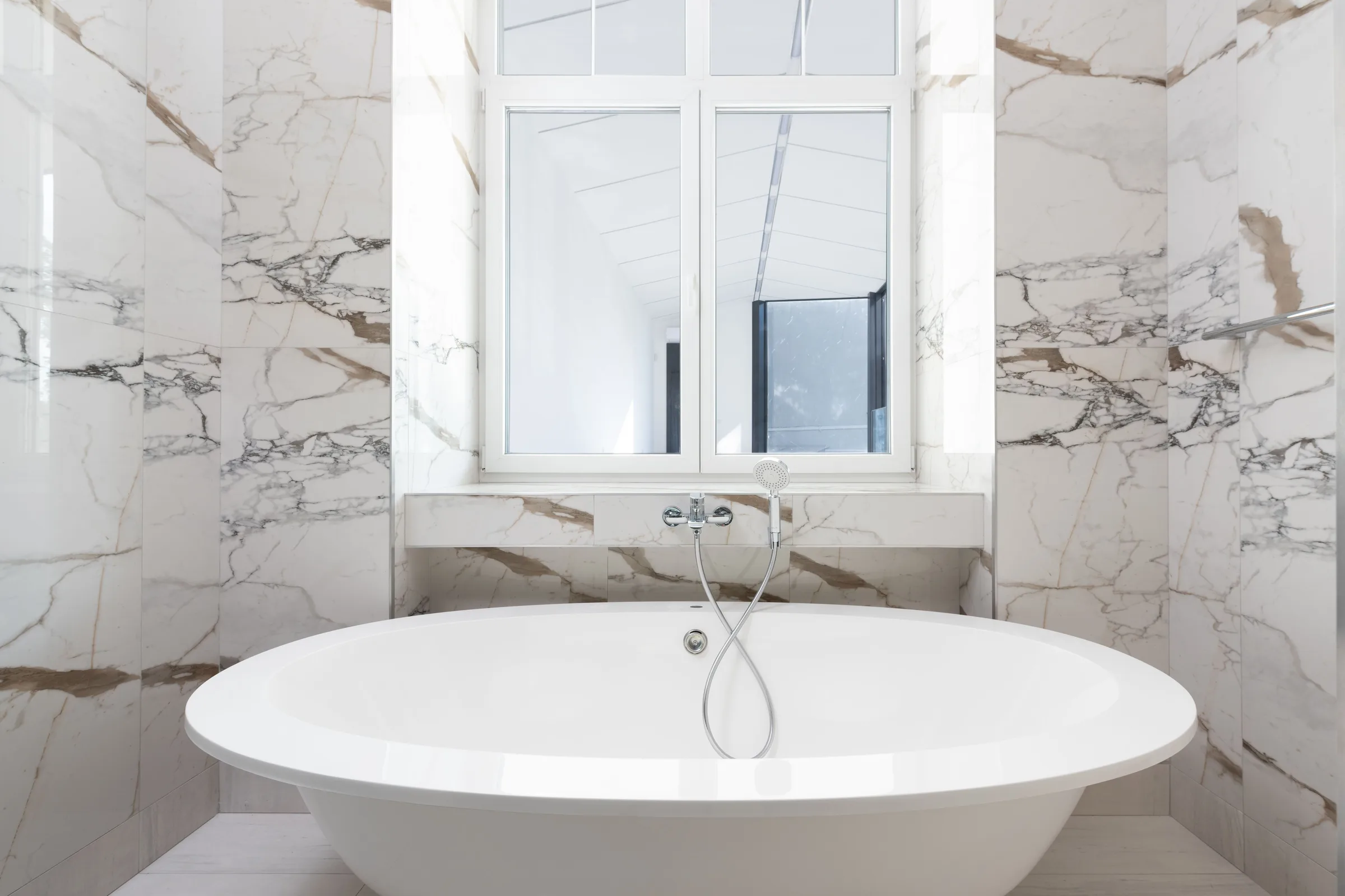 An oval freestanding tub under a white-framed window, walled in large-format white marble with gold and grey veining