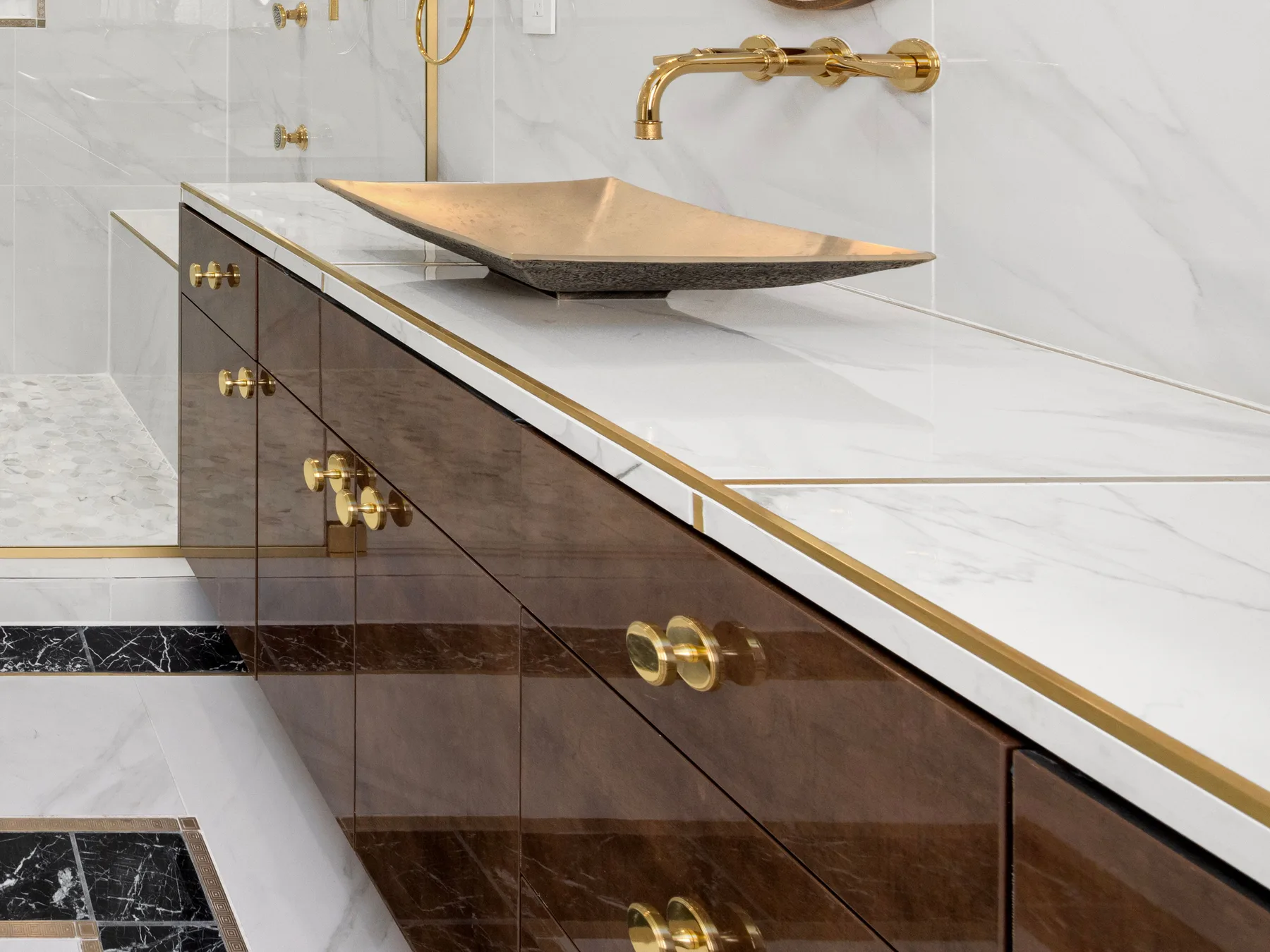 A glossy walnut vanity with round brass knobs, a white marble top edged in brass and a brass vessel basin under a wall-mounted brass faucet