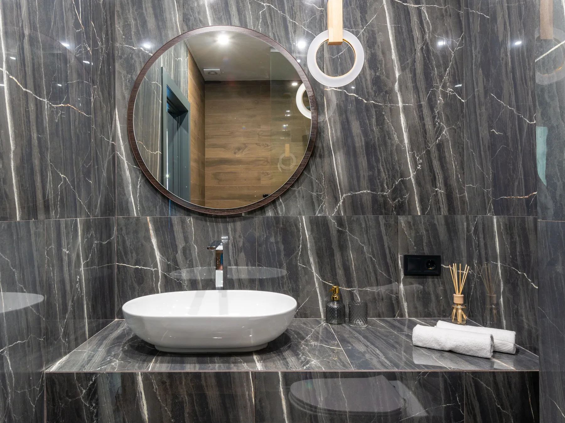 A white vessel basin on a dark marble counter against dark marble walls, under a round wood-framed mirror and a ring pendant light