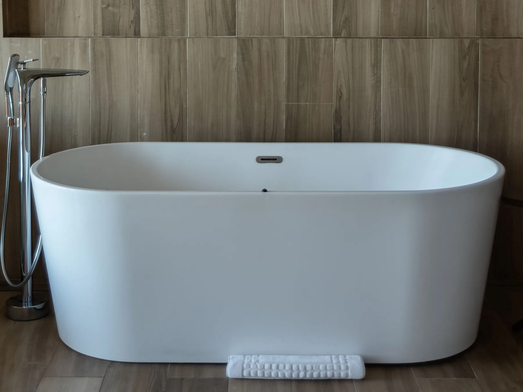 A white freestanding tub and a floor-mounted tub filler against a wood-look tiled wall