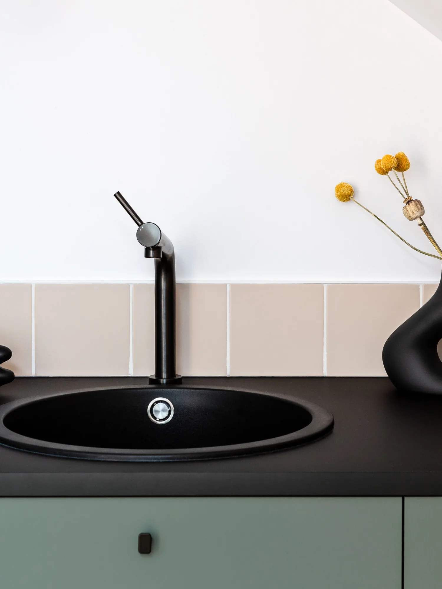 A black sink and tall faucet on a dark counter, sage-green drawers below and beige tile behind