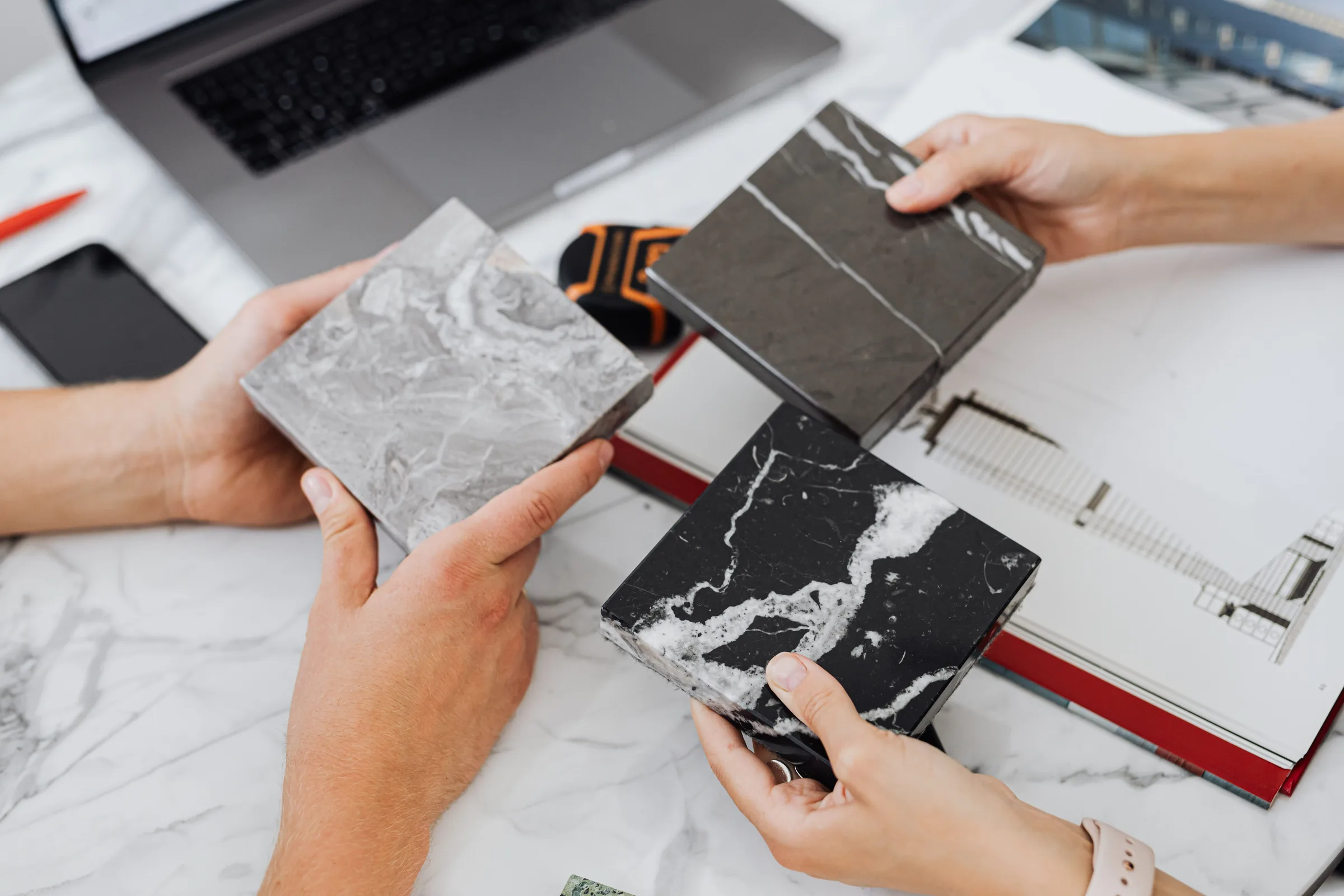 Hands comparing marble and stone samples over a catalogue and a laptop