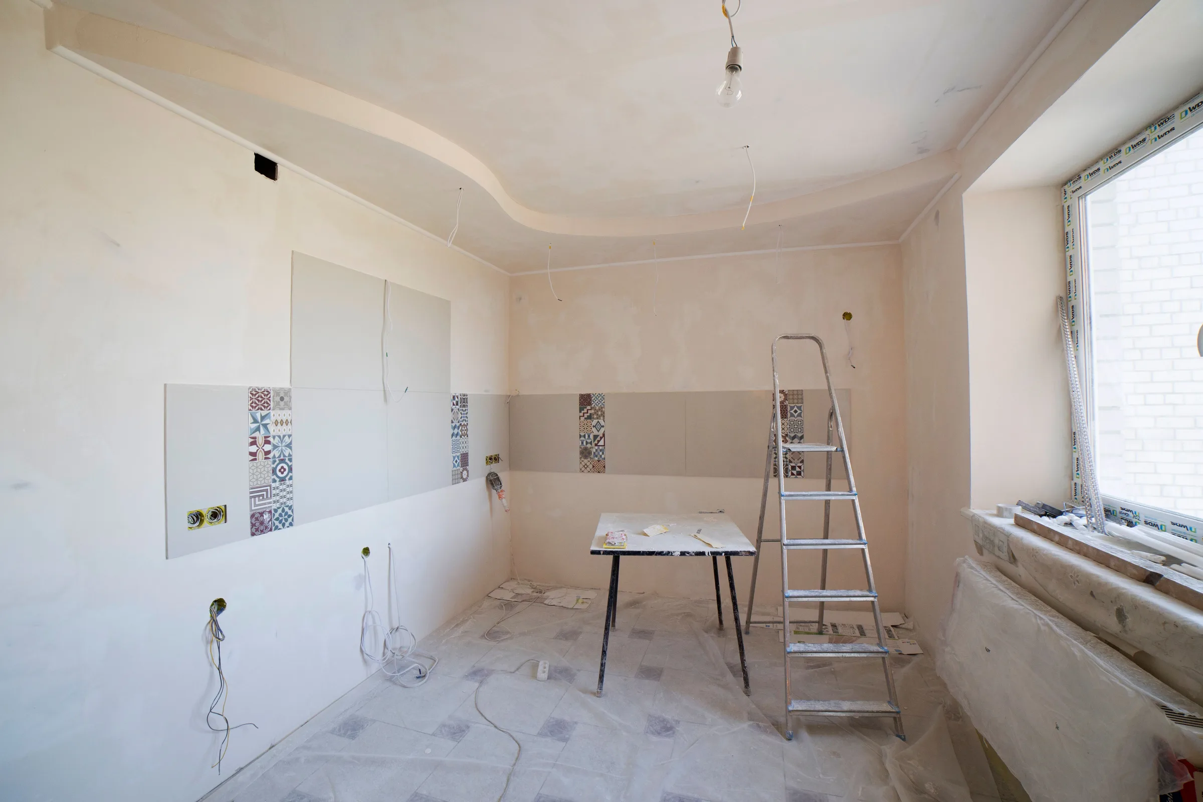 A kitchen stripped to plaster, with a stepladder, a work table and tile samples on the wall