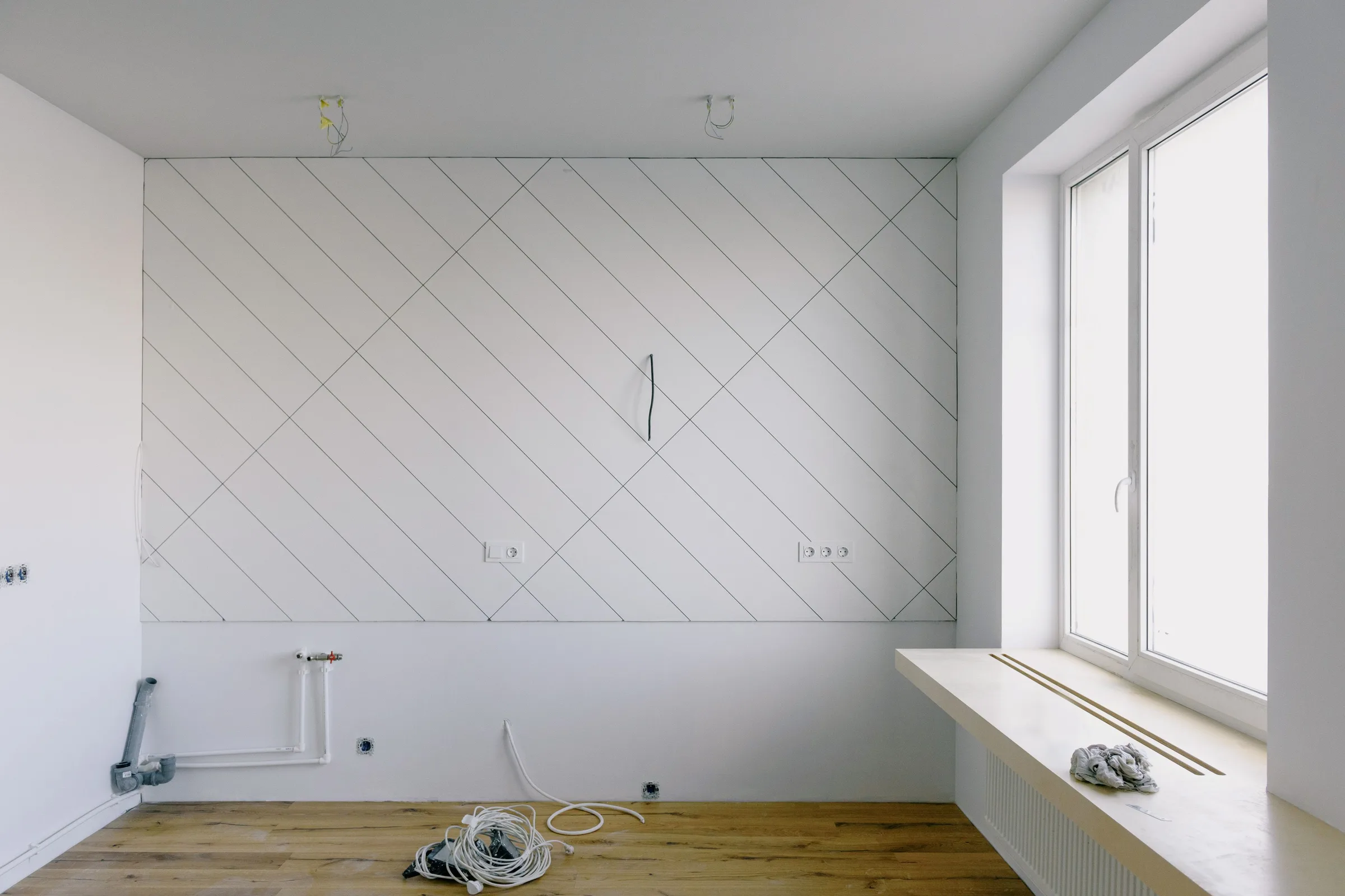 A bare room with plumbing stubs and marked tile lines on the wall, lit by a window