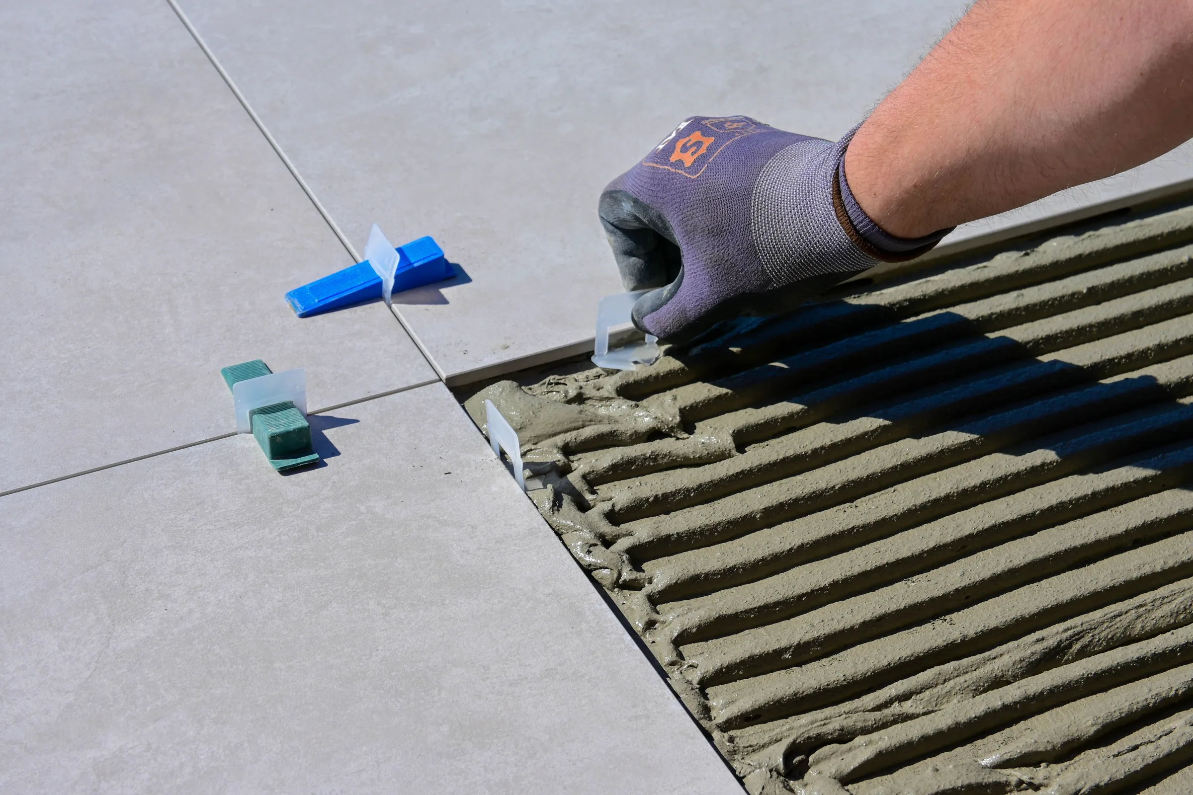 A gloved hand setting a large floor tile into combed thinset with levelling clips