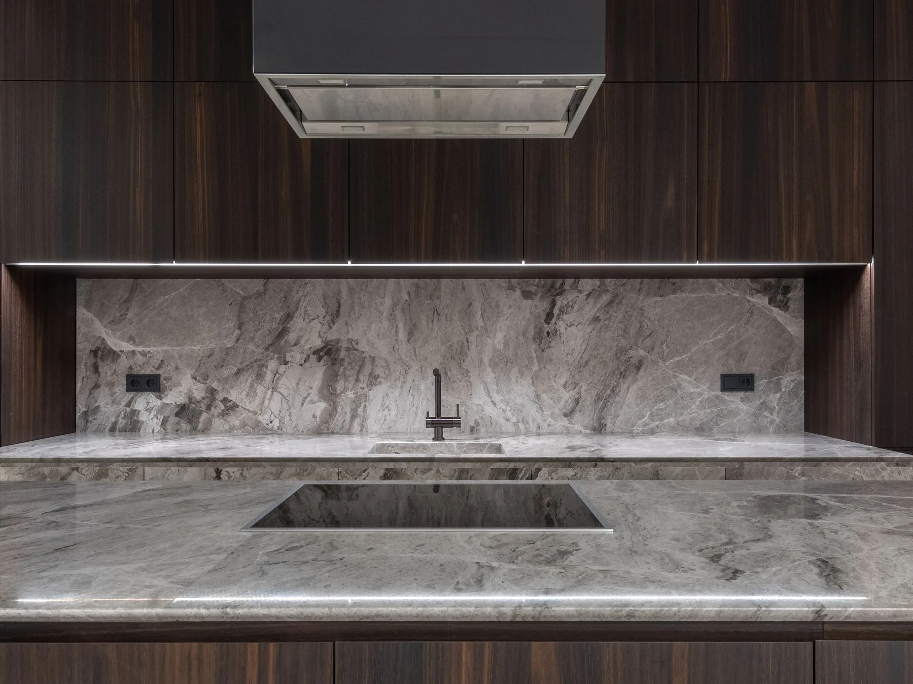 Flat dark wood cabinets around a grey stone backsplash and sink, with a black range hood above a stone island and its cooktop