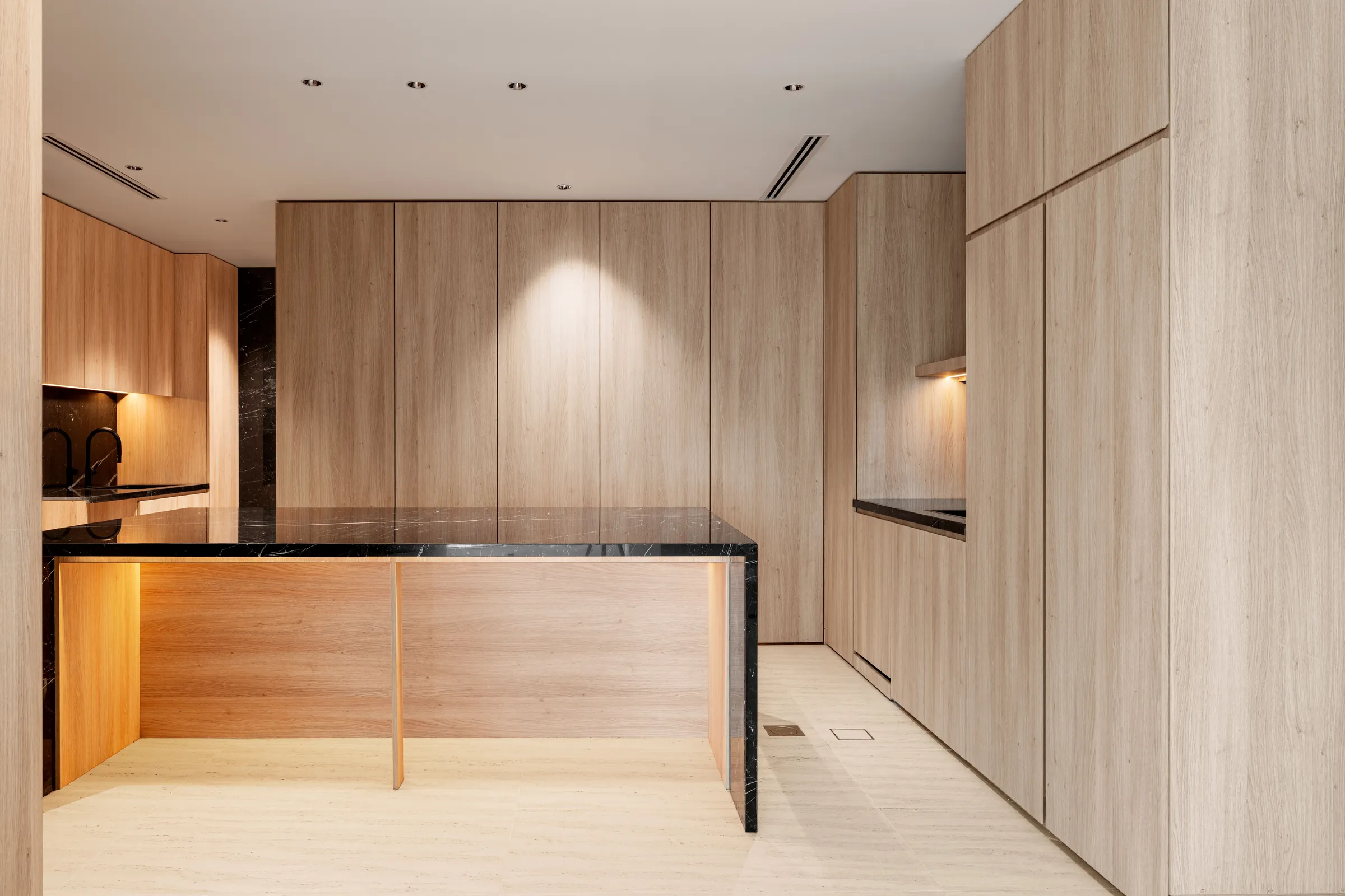 A kitchen lined in flat oak panels from floor to ceiling, with a black marble island lit from underneath and a pale stone floor