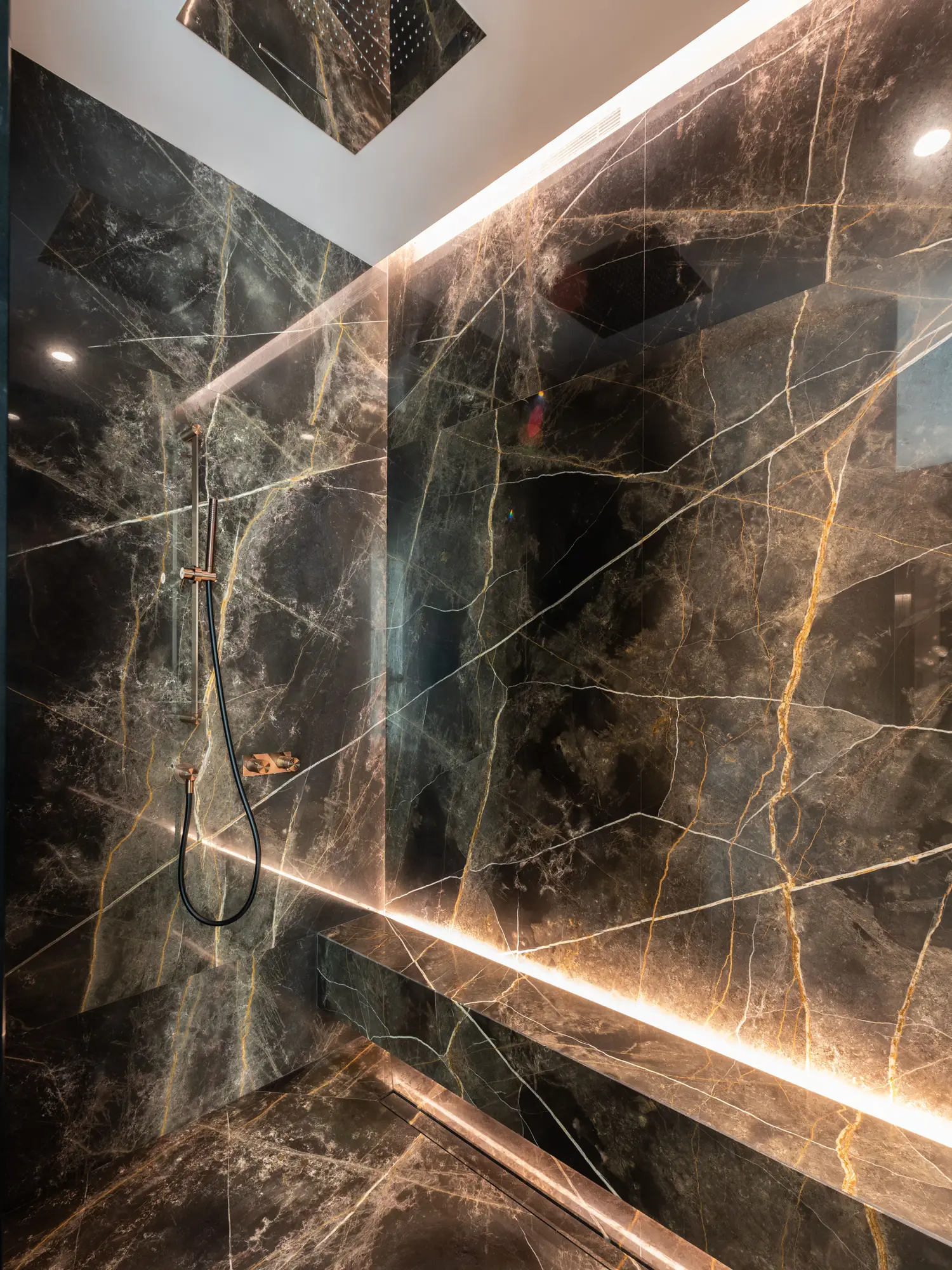 A walk-in shower walled in large dark marble slabs with gold veins, a marble bench lit by a strip light, copper fittings, a rain head in the ceiling and a linear drain