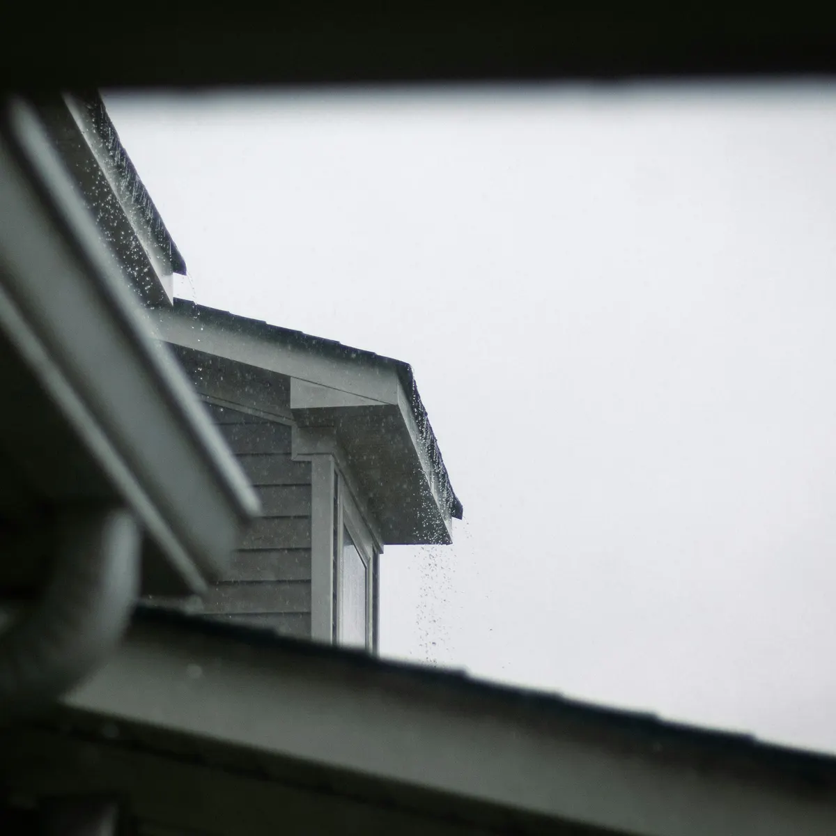 A white gutter and downspout at a roof edge against a grey sky