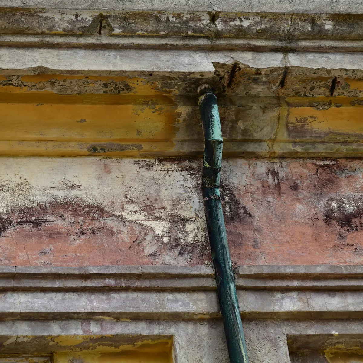 Old paint flaking off a moulded cornice beside a dark downspout
