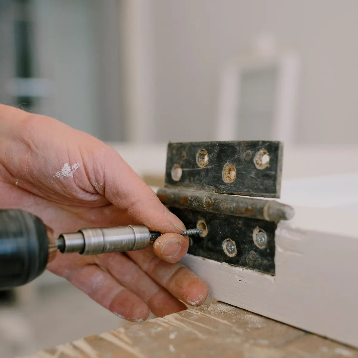A hand driving a screw into a black metal hinge with a drill