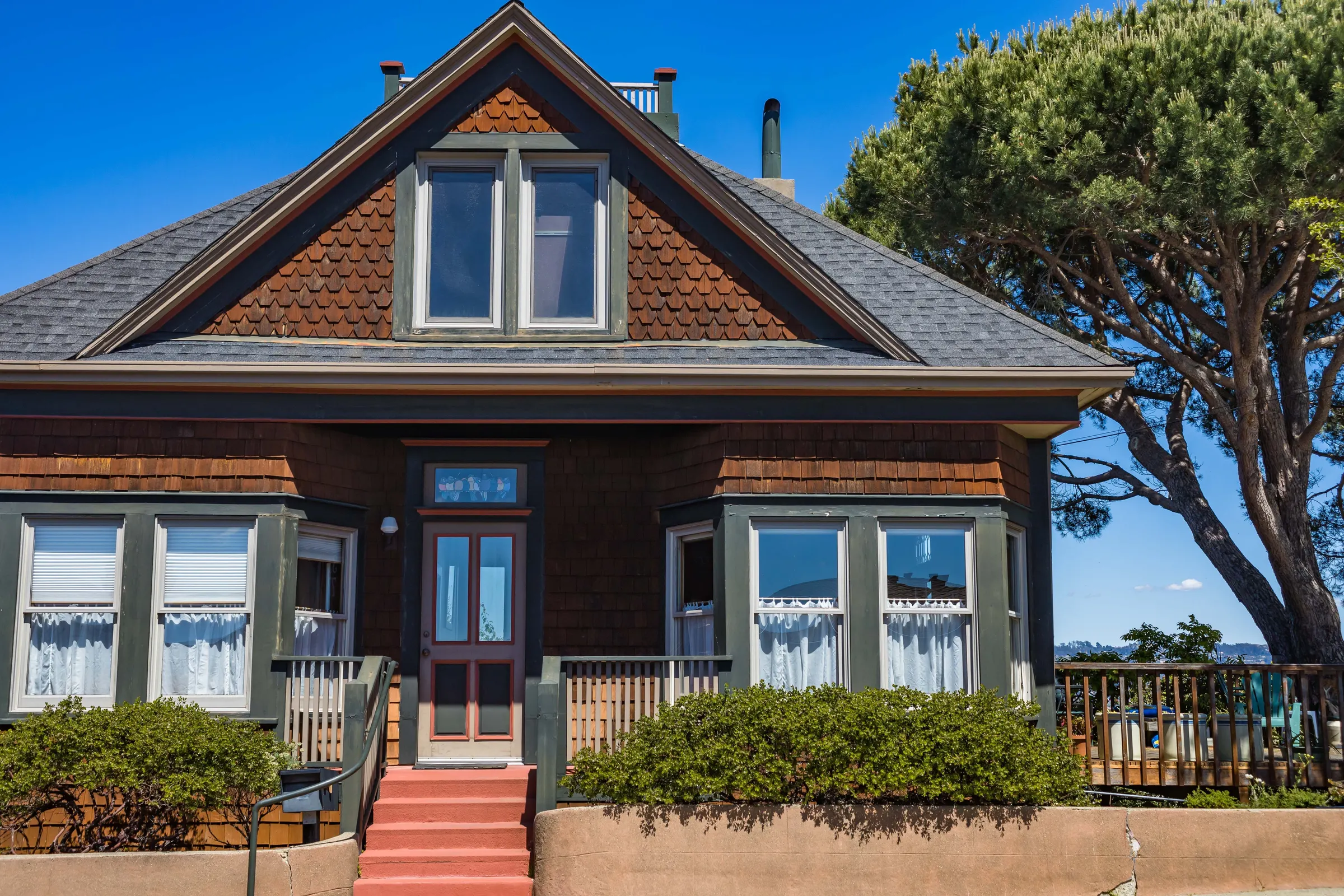 A brown shingled house with a steep gable, two bay windows, red front steps and a pine beside it