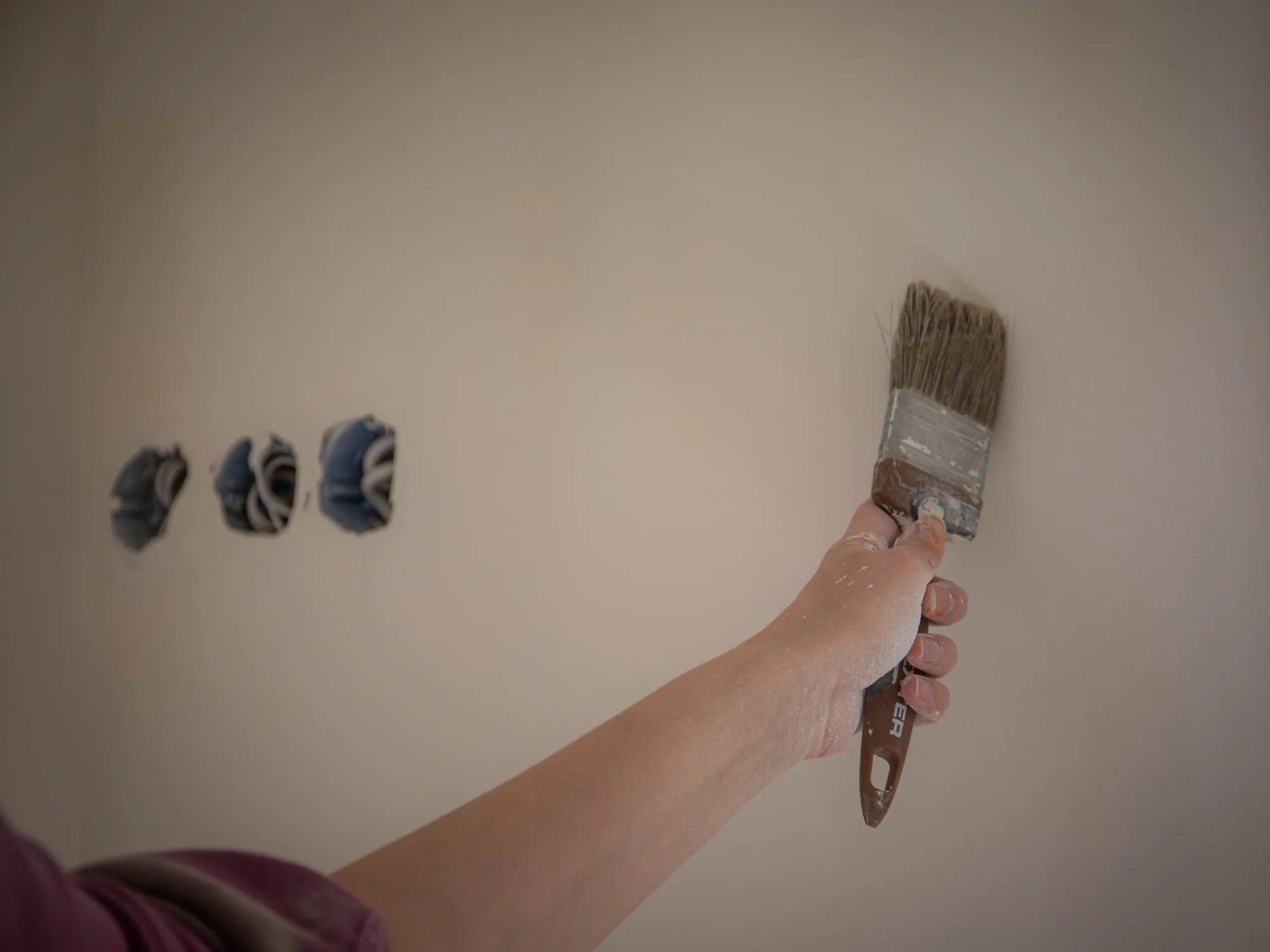A hand with a paintbrush touching up a pale interior wall