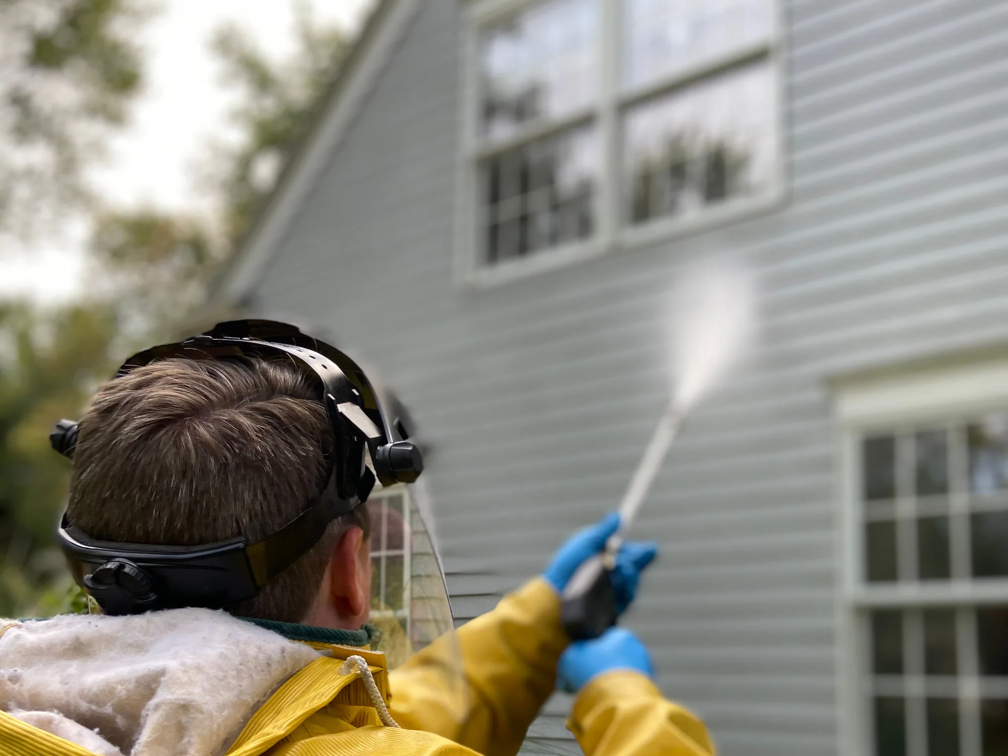 A person in a yellow rain jacket spraying grey house siding, seen from behind