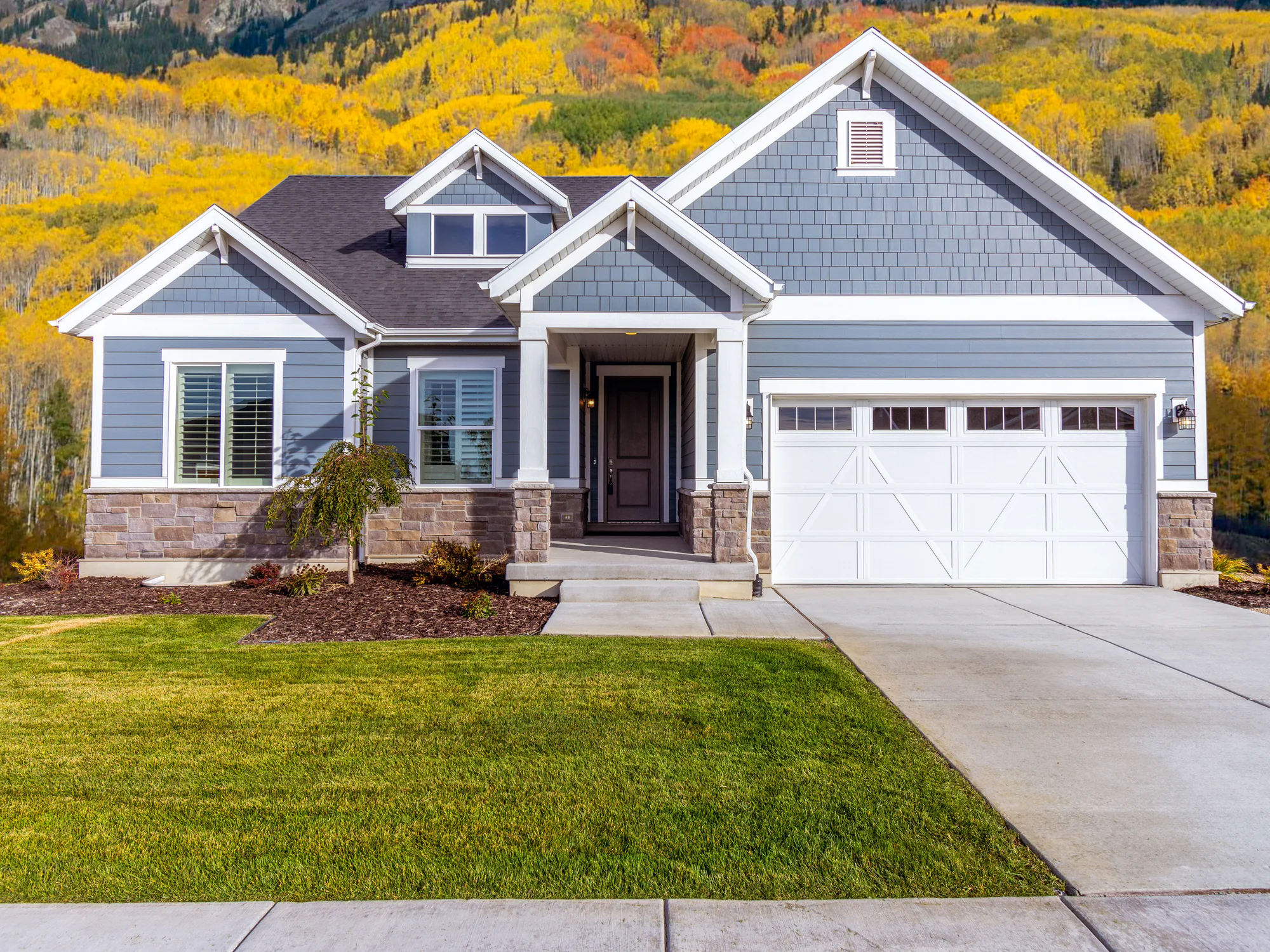 A blue-grey house with a mown lawn below a hillside of autumn trees