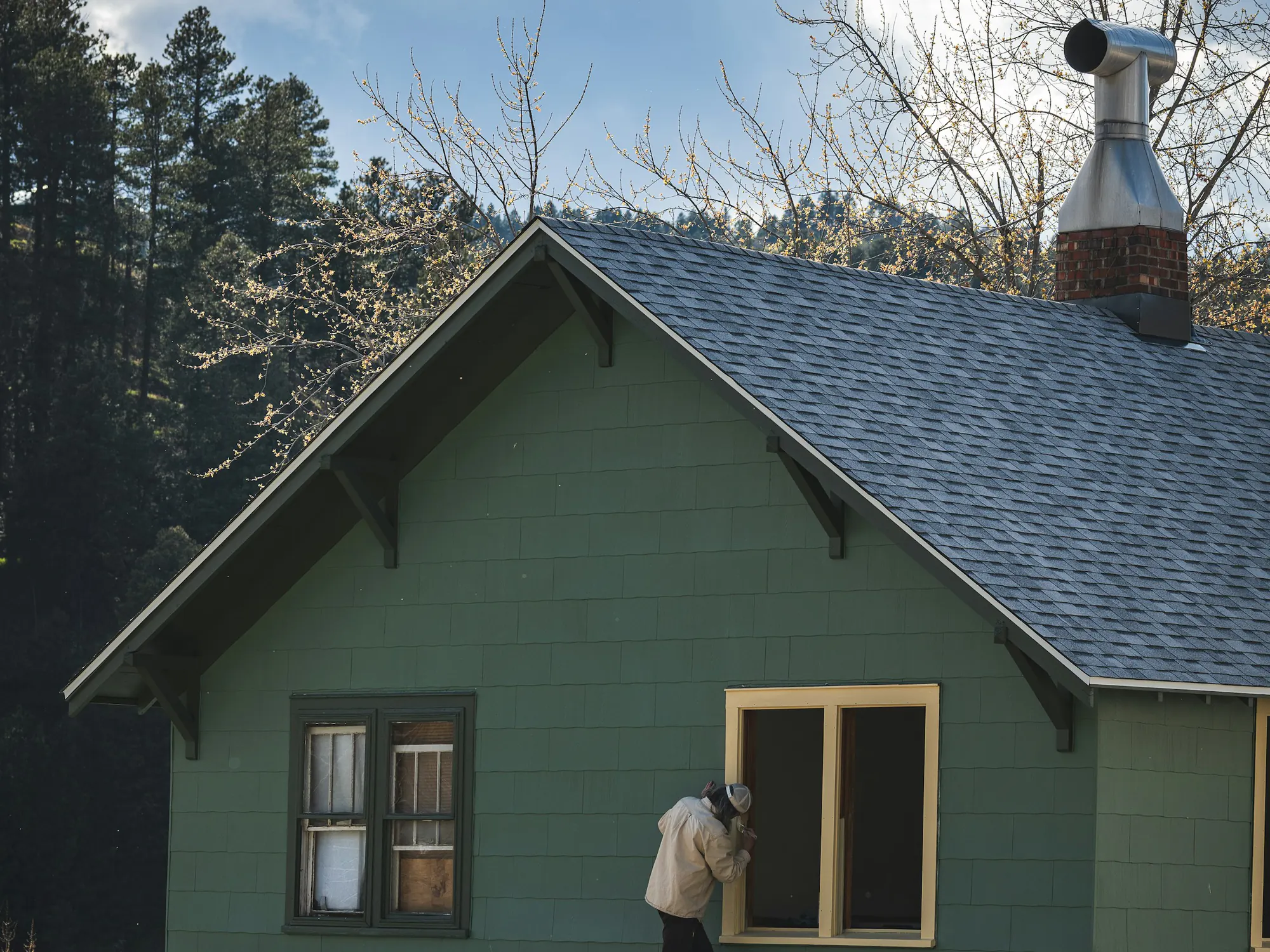 A painter on a porch roof painting the trim of a window on a green shingled Craftsman house