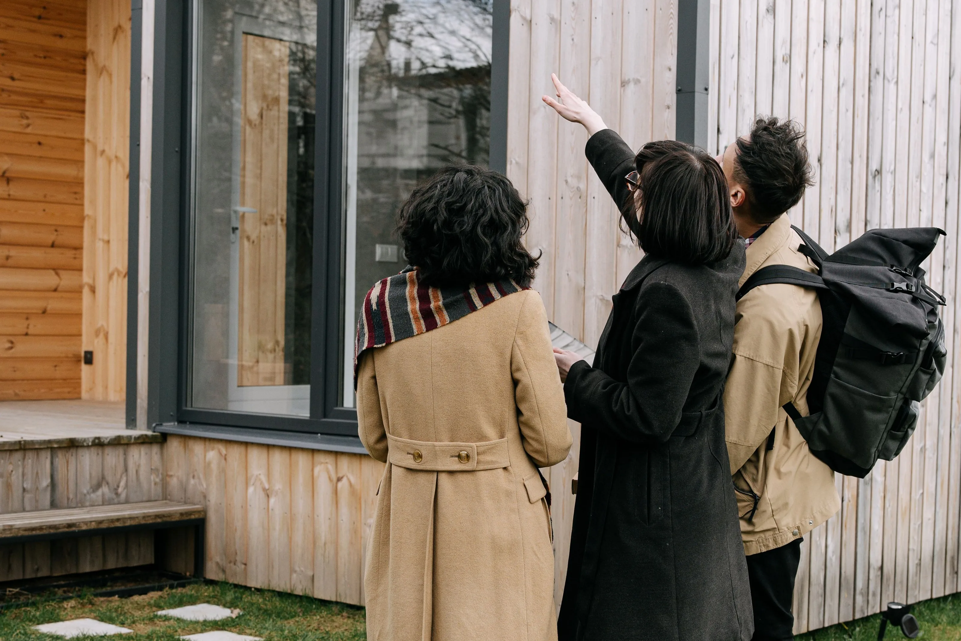 Three people seen from behind on a lawn, one pointing up at the tall windows of a wood-clad house
