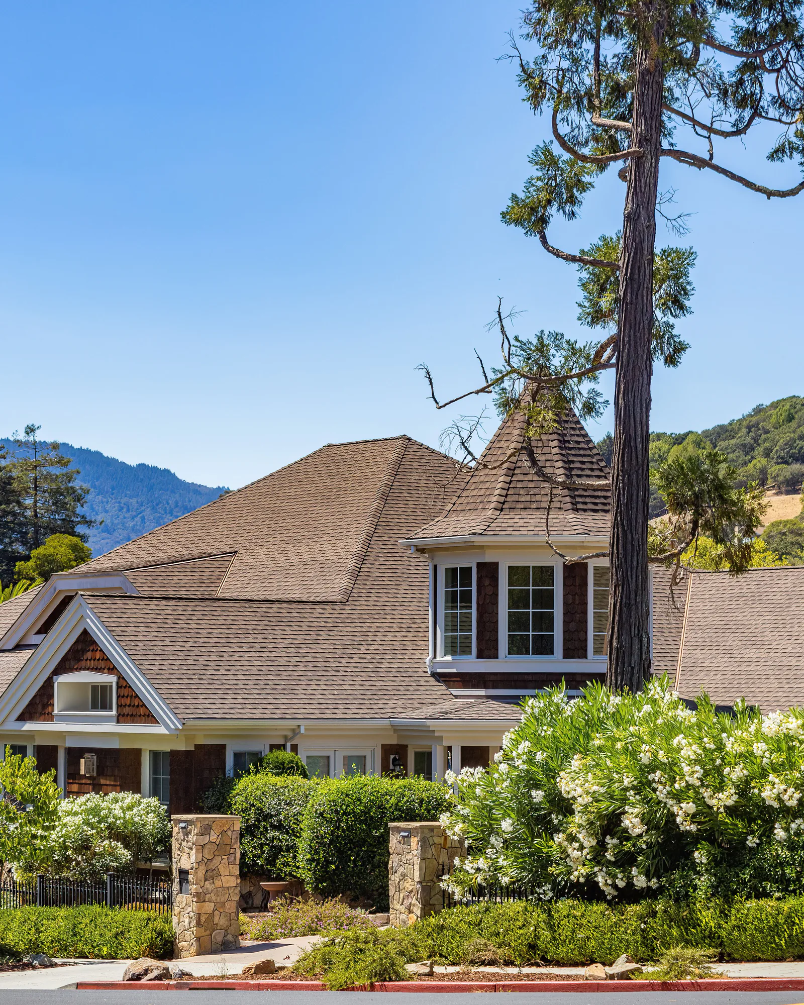 A shingled house among trimmed shrubs and tall pines below a wooded mountain