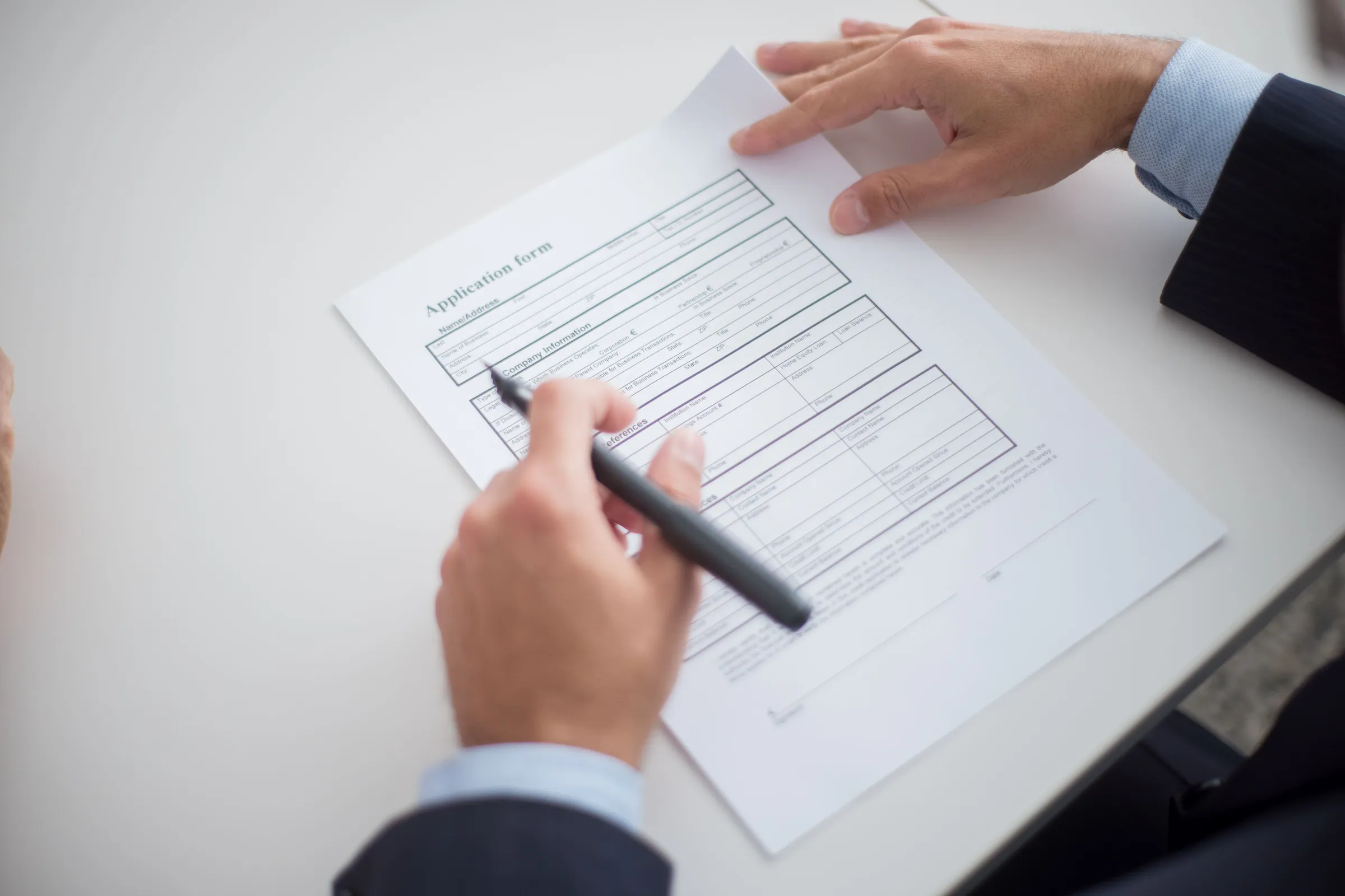 Two hands over a printed application form on a white desk, one holding a pen