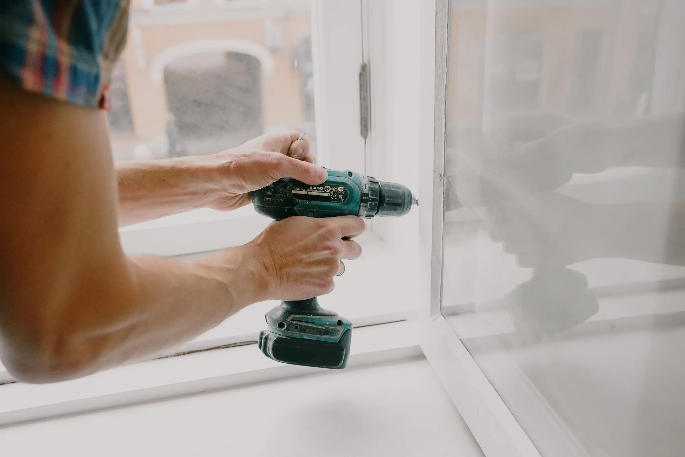 Two hands driving a screw into a white window frame with a cordless drill
