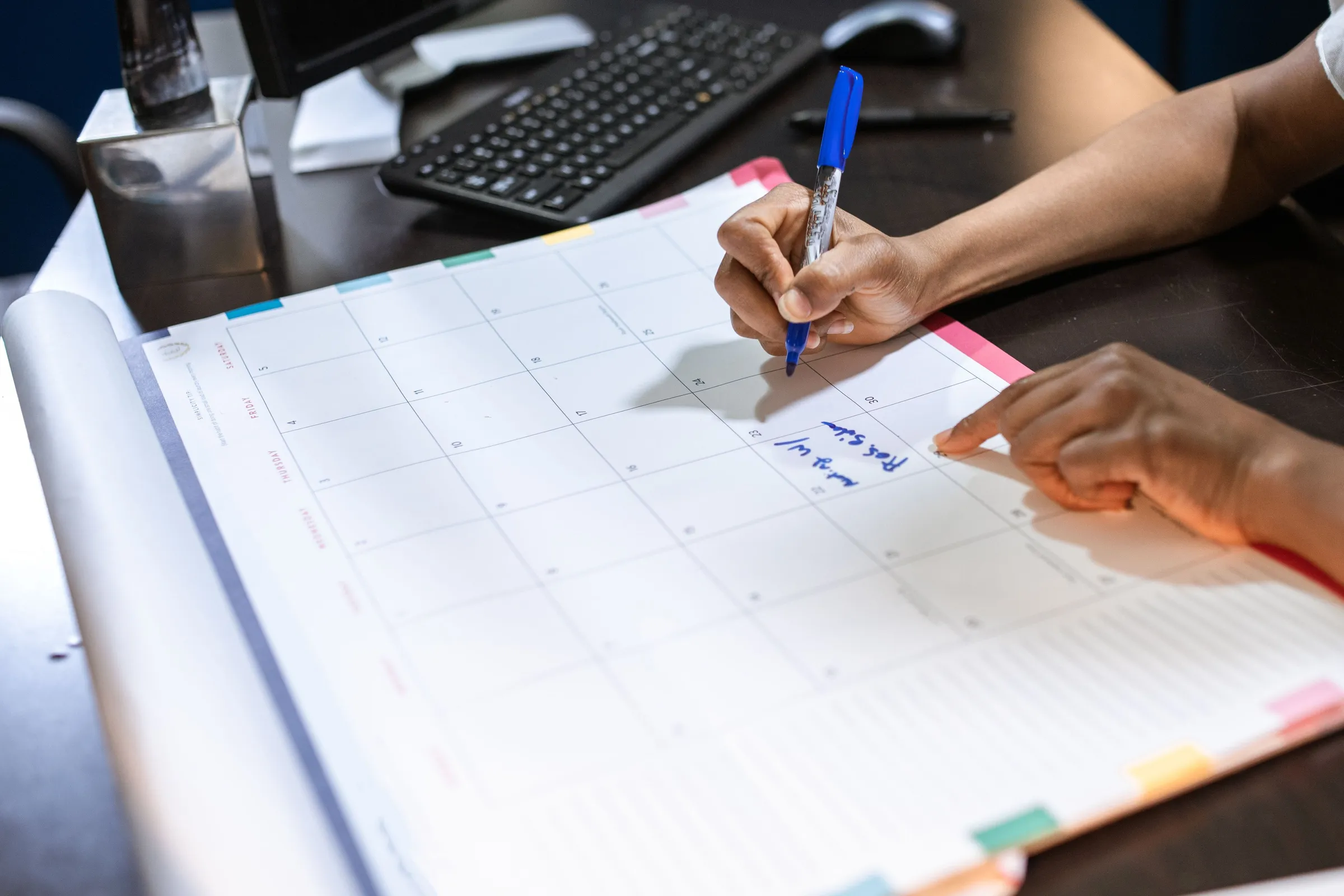 Hands marking a date on a large desk calendar with a blue pen, a keyboard behind