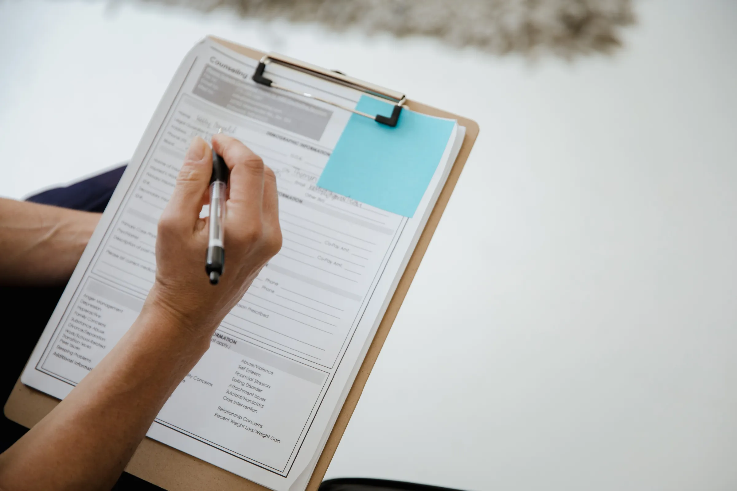 A hand with a pen over a printed checklist on a clipboard, a teal sticky note at its corner