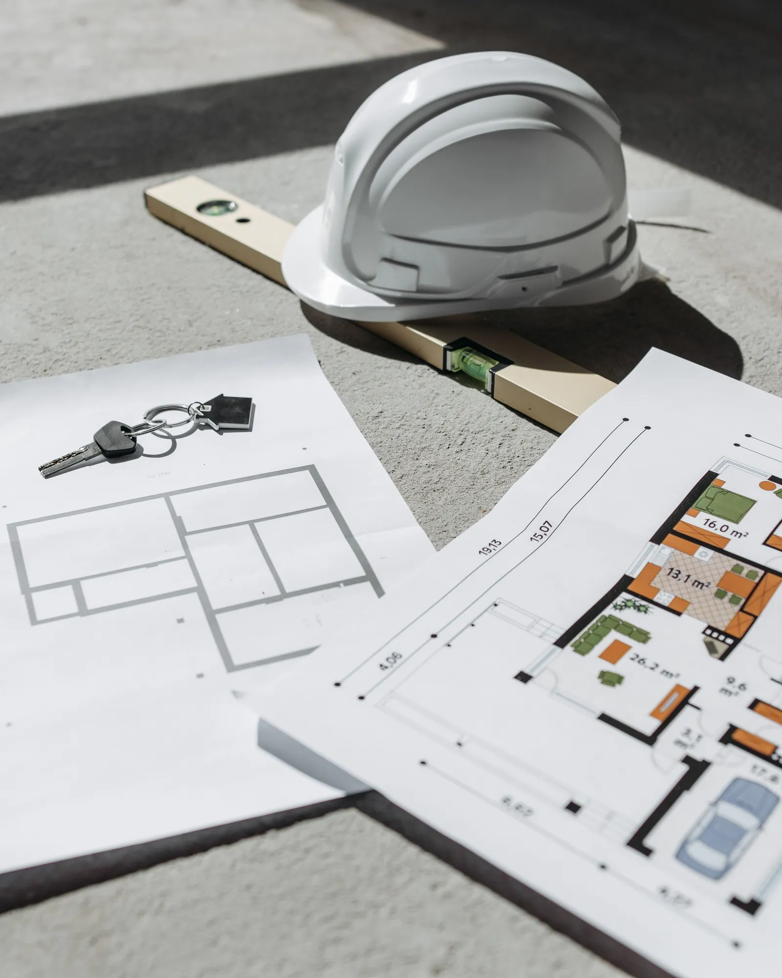 Two floor plan sheets, a set of keys, a spirit level and a white hard hat on a sunlit concrete floor