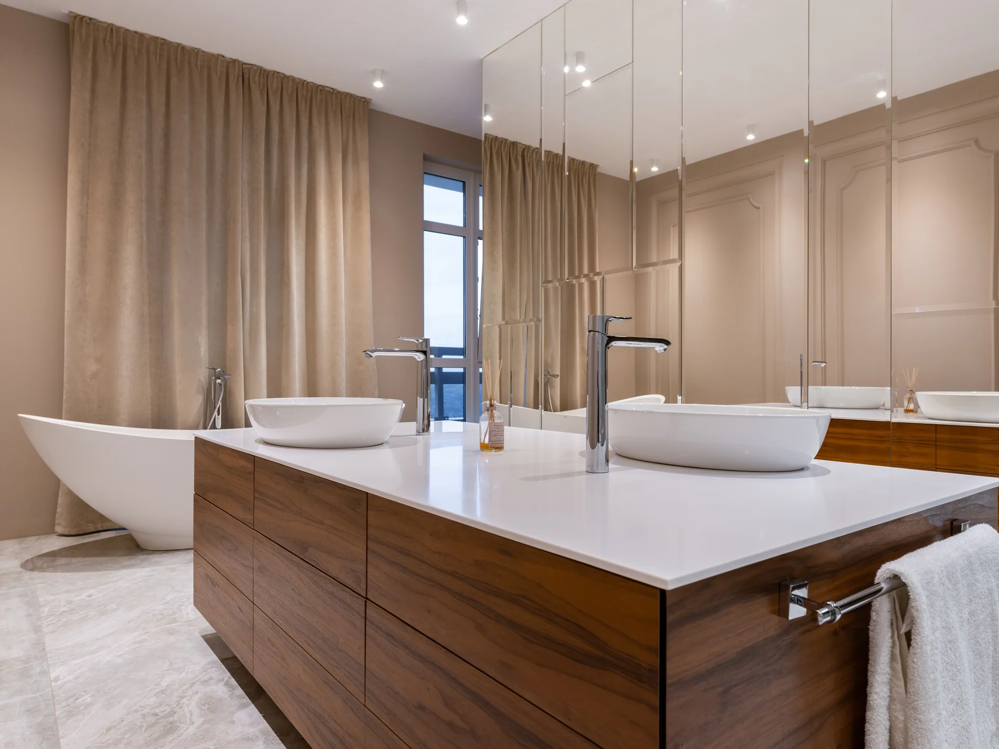 A walnut slab-front vanity island with a white top and two vessel basins, a freestanding tub and floor-length taupe curtains behind
