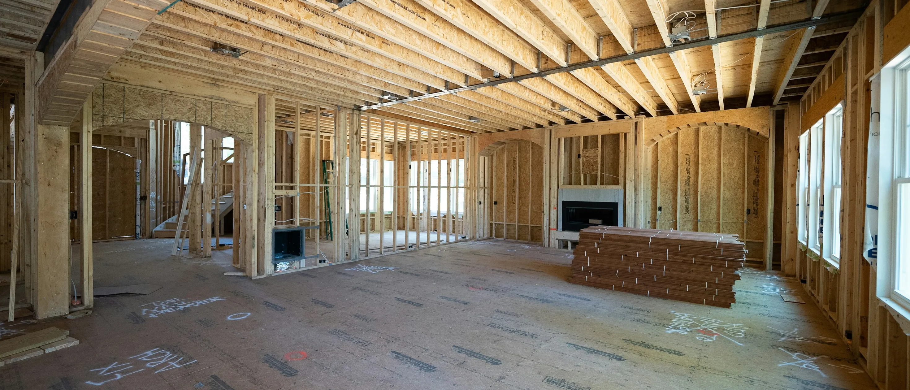 A large room opened to its wood framing and ceiling joists mid-remodel, the subfloor marked up