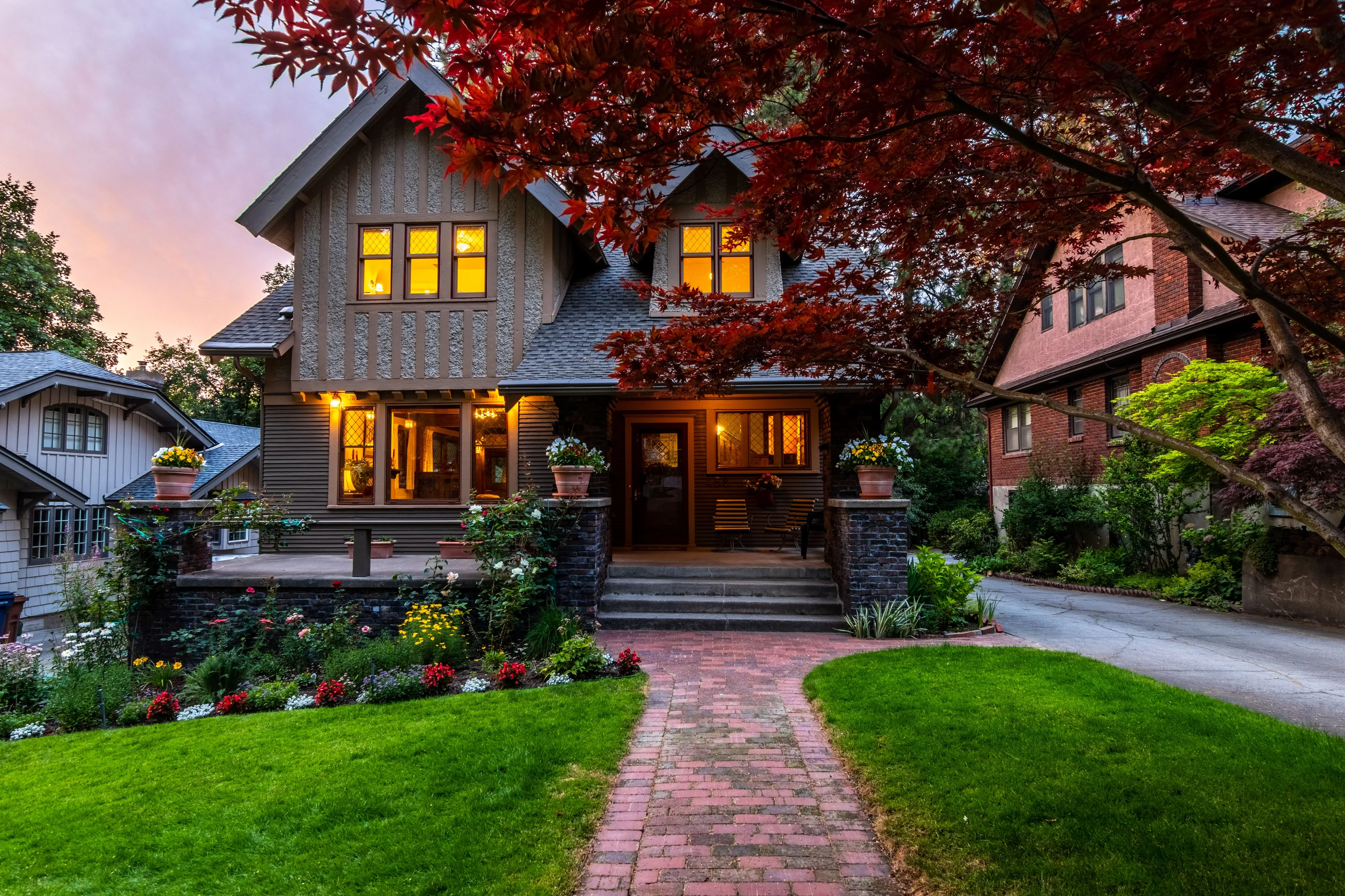 A Craftsman house with a lit porch and a brick path at twilight, under a red maple